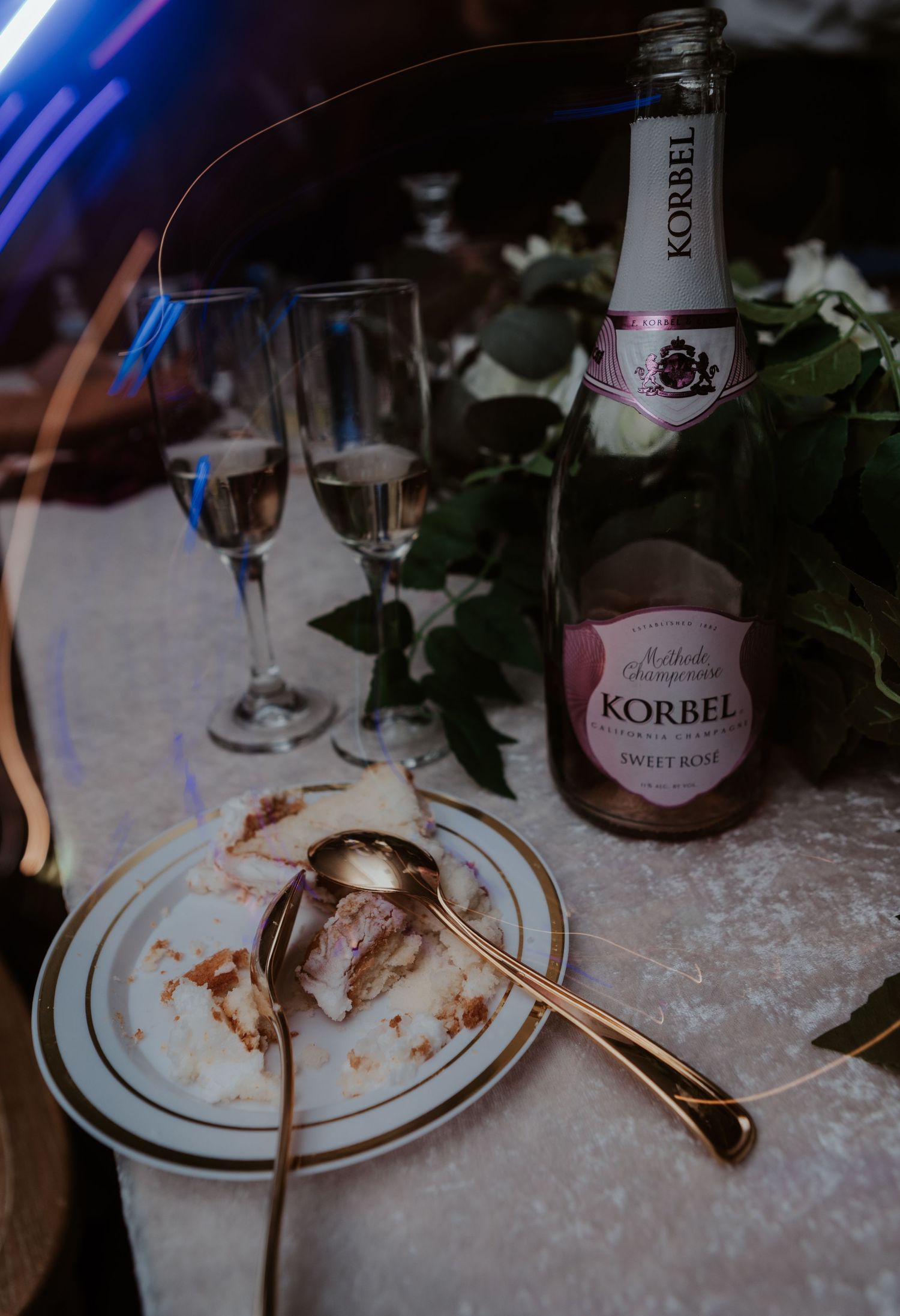 A dessert plate with leftover crumbs beside a bottle of Korbel Sweet Rose champagne and two flutes on a white tablecloth.