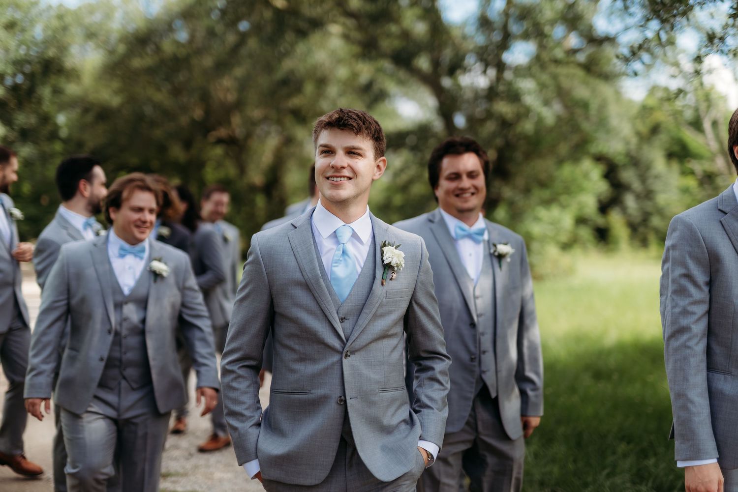 Groomsmen wearing light gray suits with blue ties pose together outdoors on a sunny day.