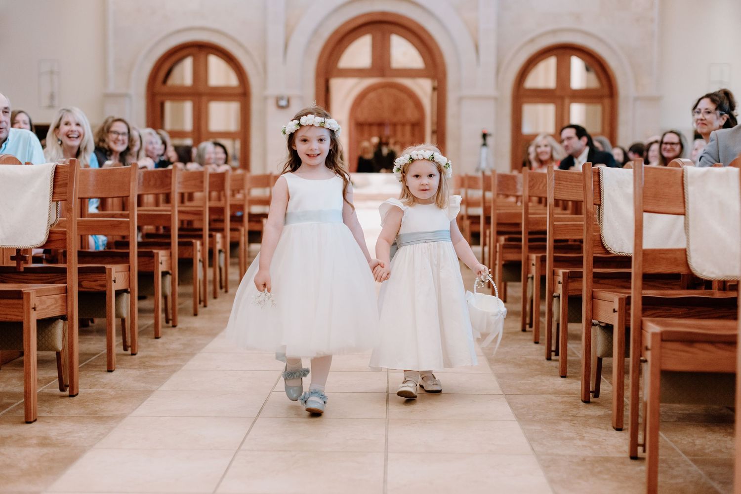 Two flower girls in white dresses walk down church aisle holding hands as guests watch from wooden pews.