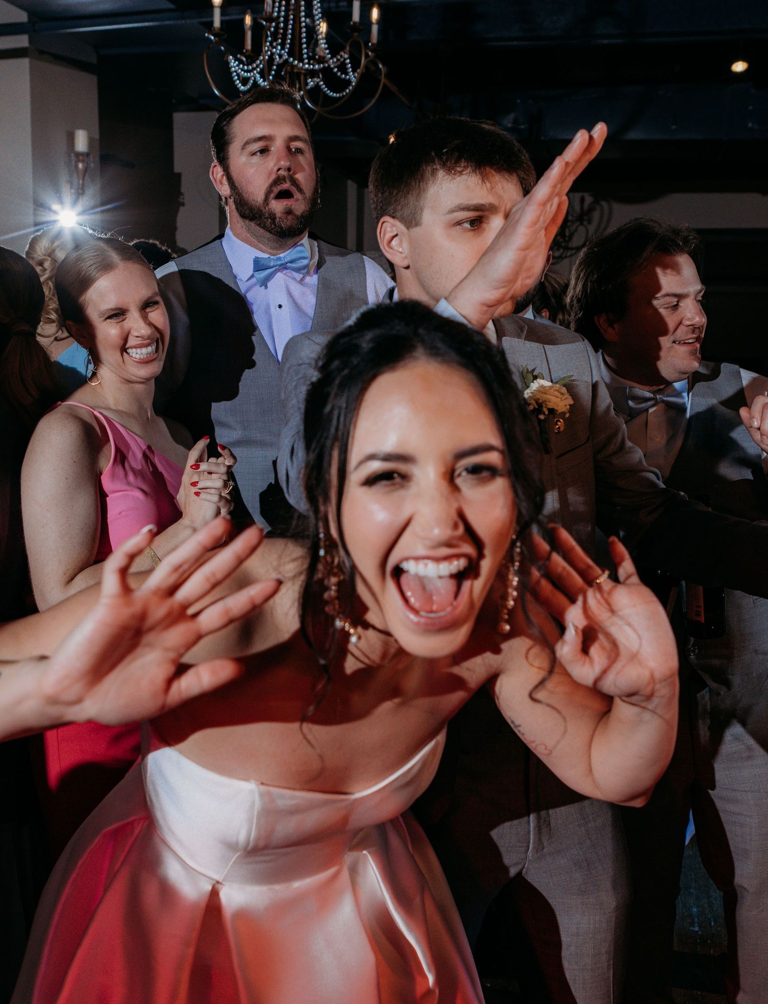 Wedding guests dancing and celebrating with joyful expressions at a reception party under chandeliers.