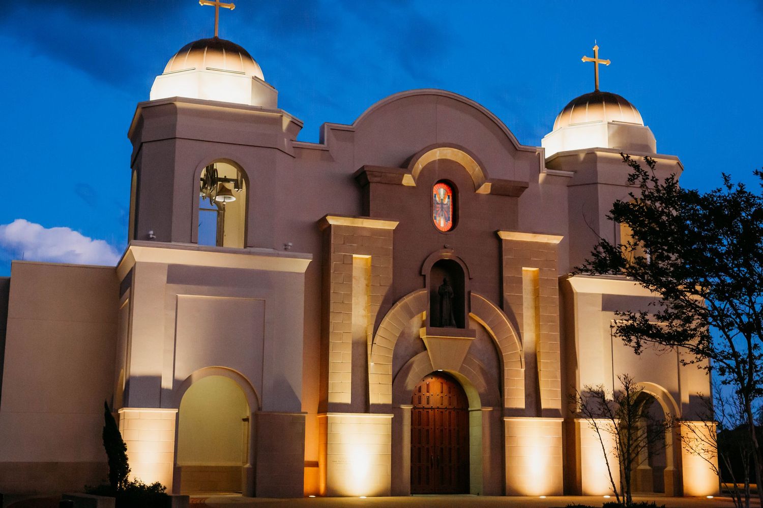 Spanish colonial style mission church with white stucco walls and golden domes illuminated at dusk against blue sky.