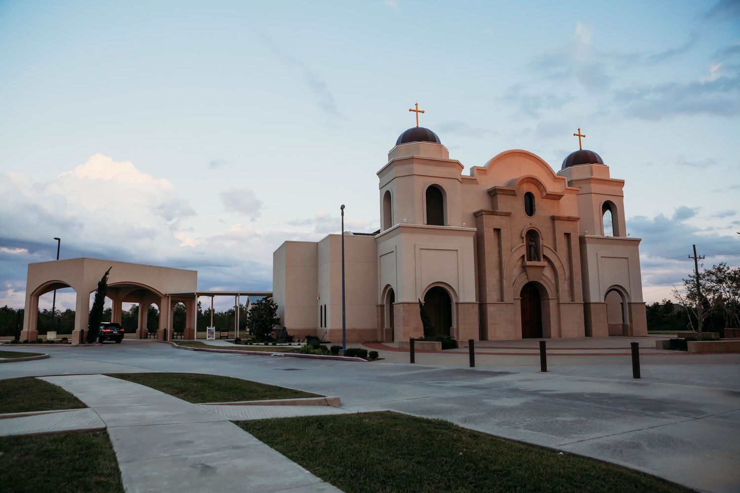 Spanish colonial style Catholic church with twin bell towers and domes stands at dusk against cloudy sky in Southwest design.