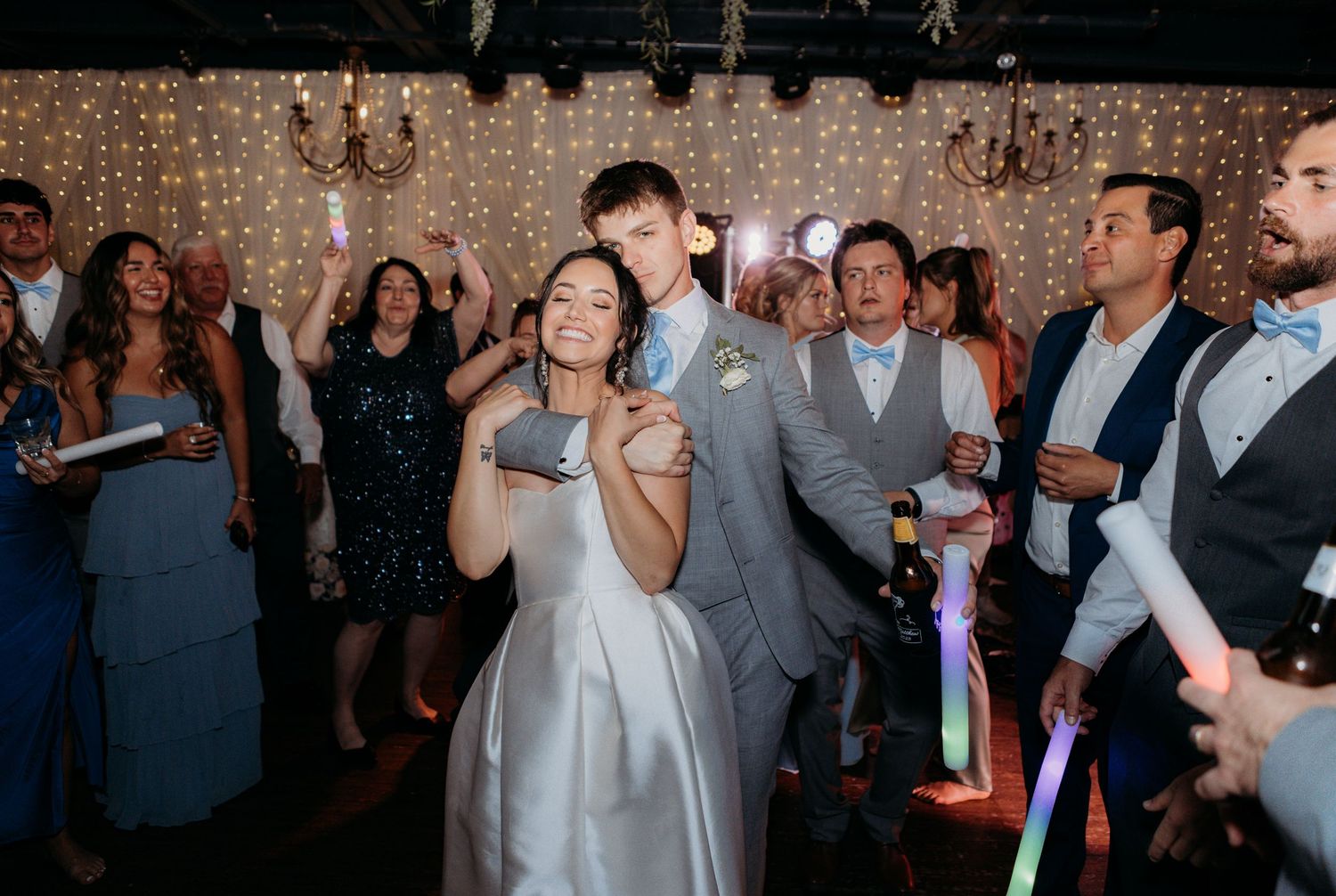 Wedding celebration with guests dancing and holding glow sticks under sparkling lights.