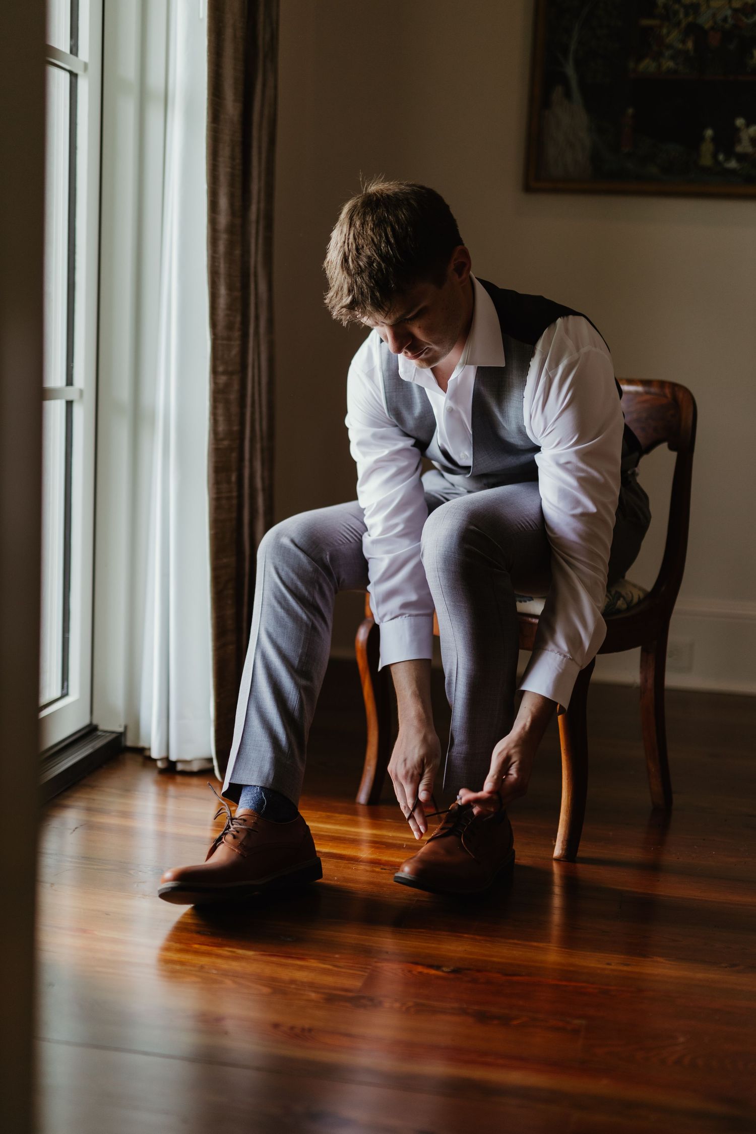 Elegant figure in white shirt and vest sits by window on wooden chair tying brown leather dress shoes on hardwood floor.