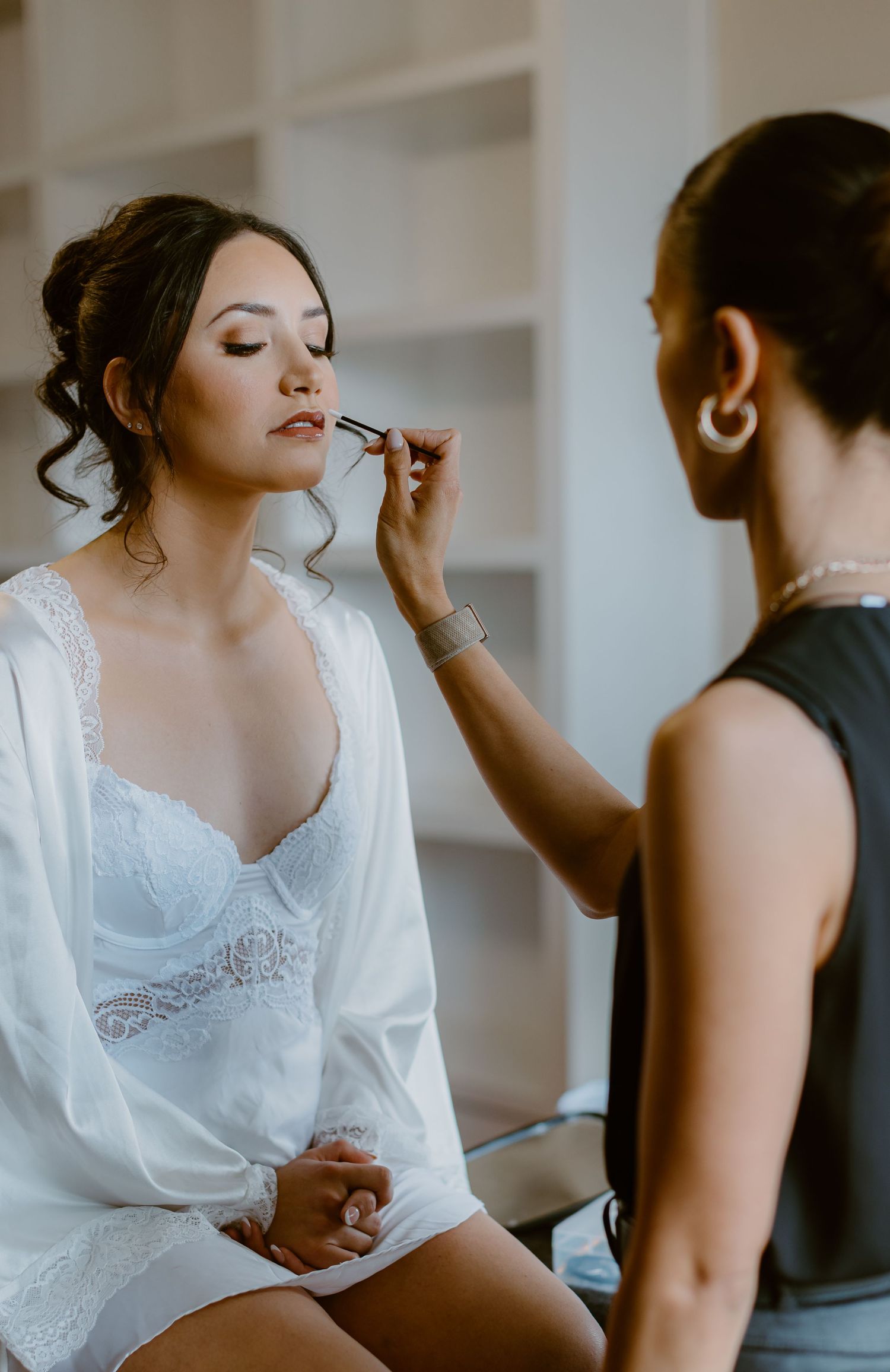 A makeup artist applies product to a person wearing a white lace robe during wedding day preparations.