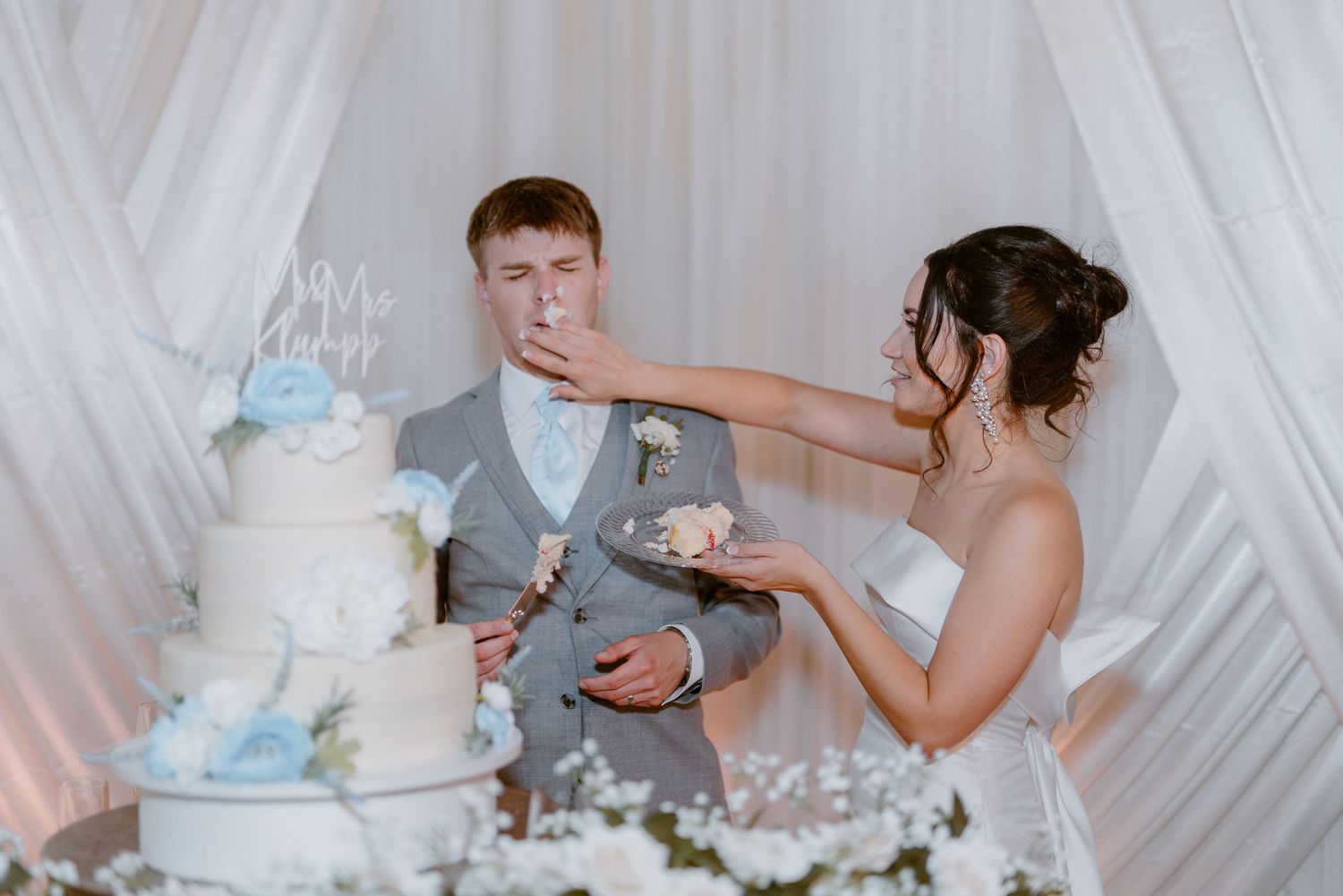 A couple shares a playful cake-feeding moment beside their elegant white and blue tiered wedding cake.