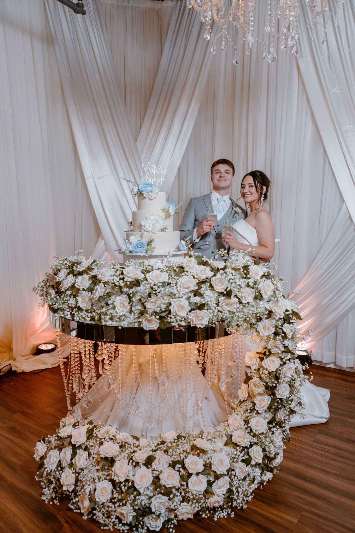 Elegant wedding cake display with cascading white floral arrangements on a suspended lit table with draped fabric backdrop.