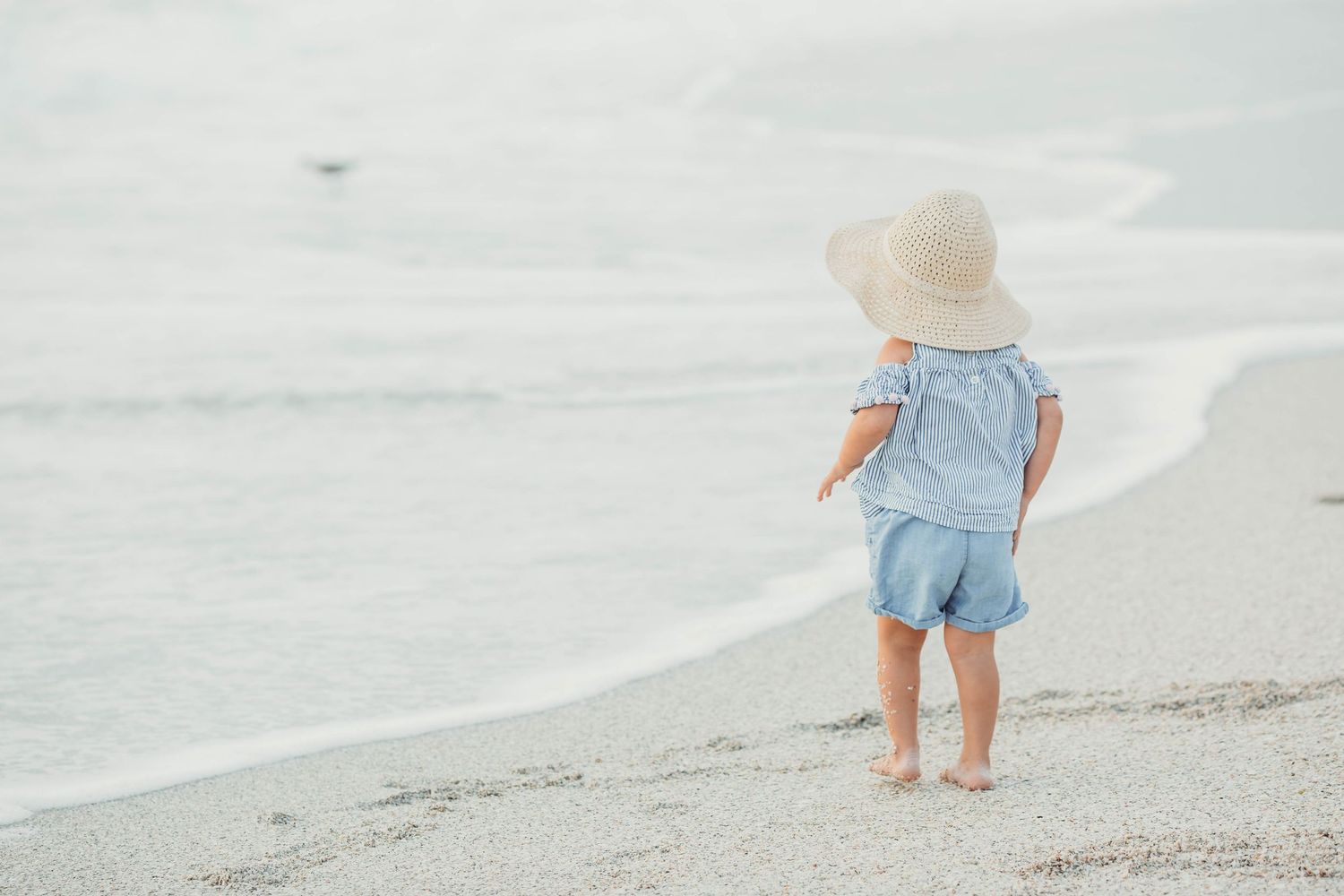 Small child in light blue overalls and sun hat stands barefoot on a peaceful beach near the ocean waves.