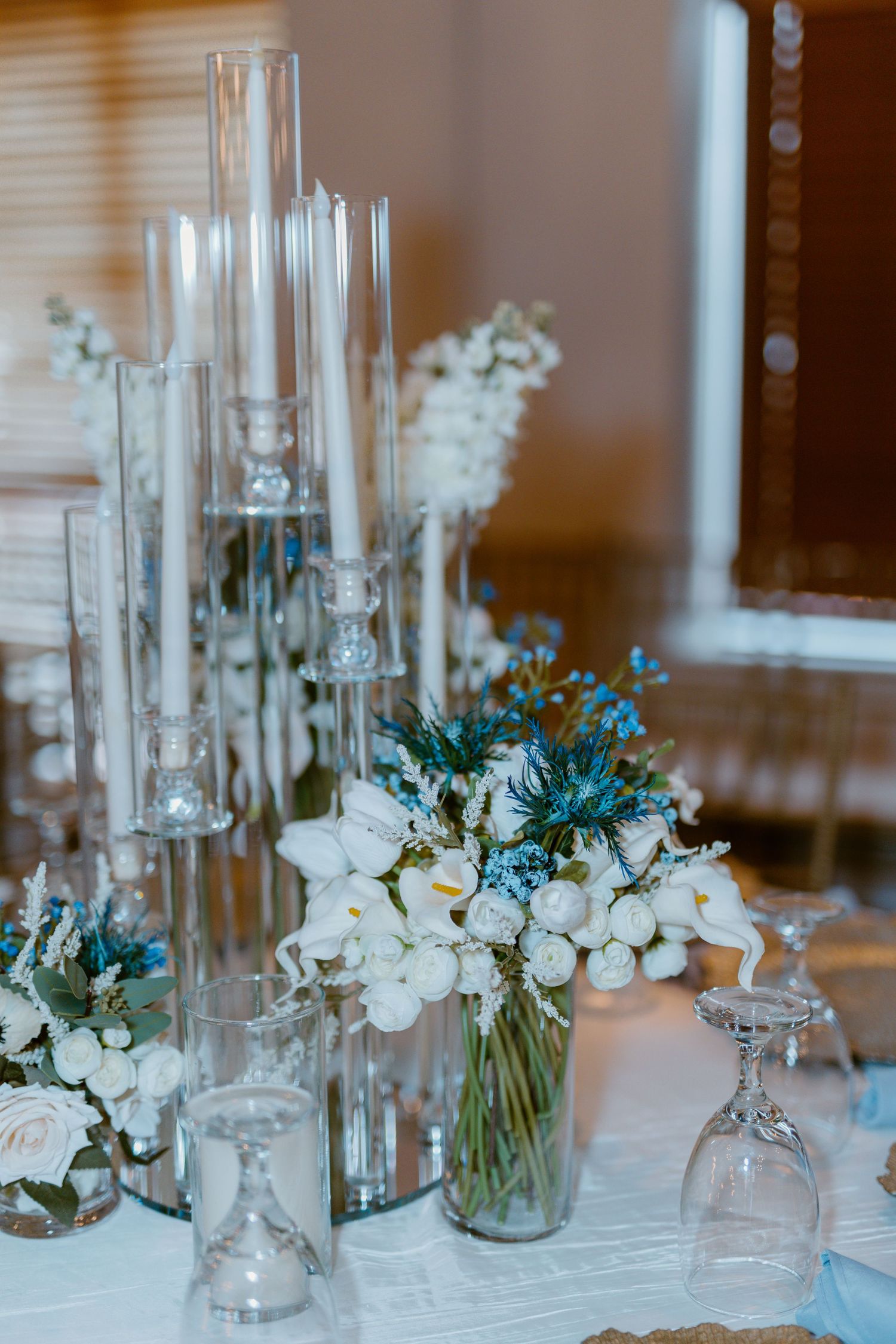 Elegant wedding table centerpiece with white flowers, blue thistles and tall glass candle holders on white tablecloth.