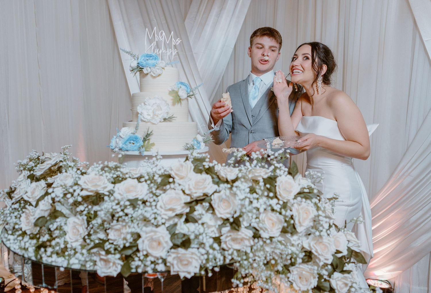 Newlywed couple stands at their wedding reception table adorned with white floral arrangements and a tiered cake.