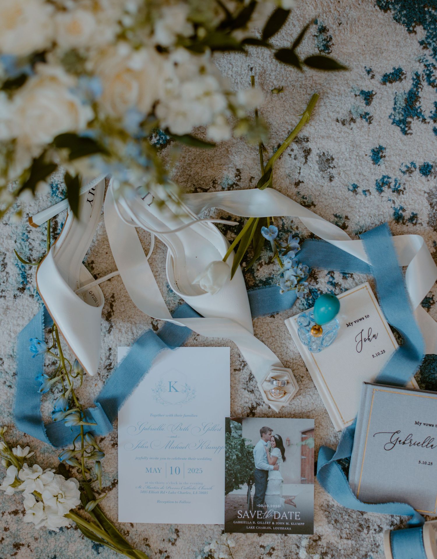 Wedding stationery arranged with white ribbons, flowers and invitation cards in soft blue and white tones on a textured surface.