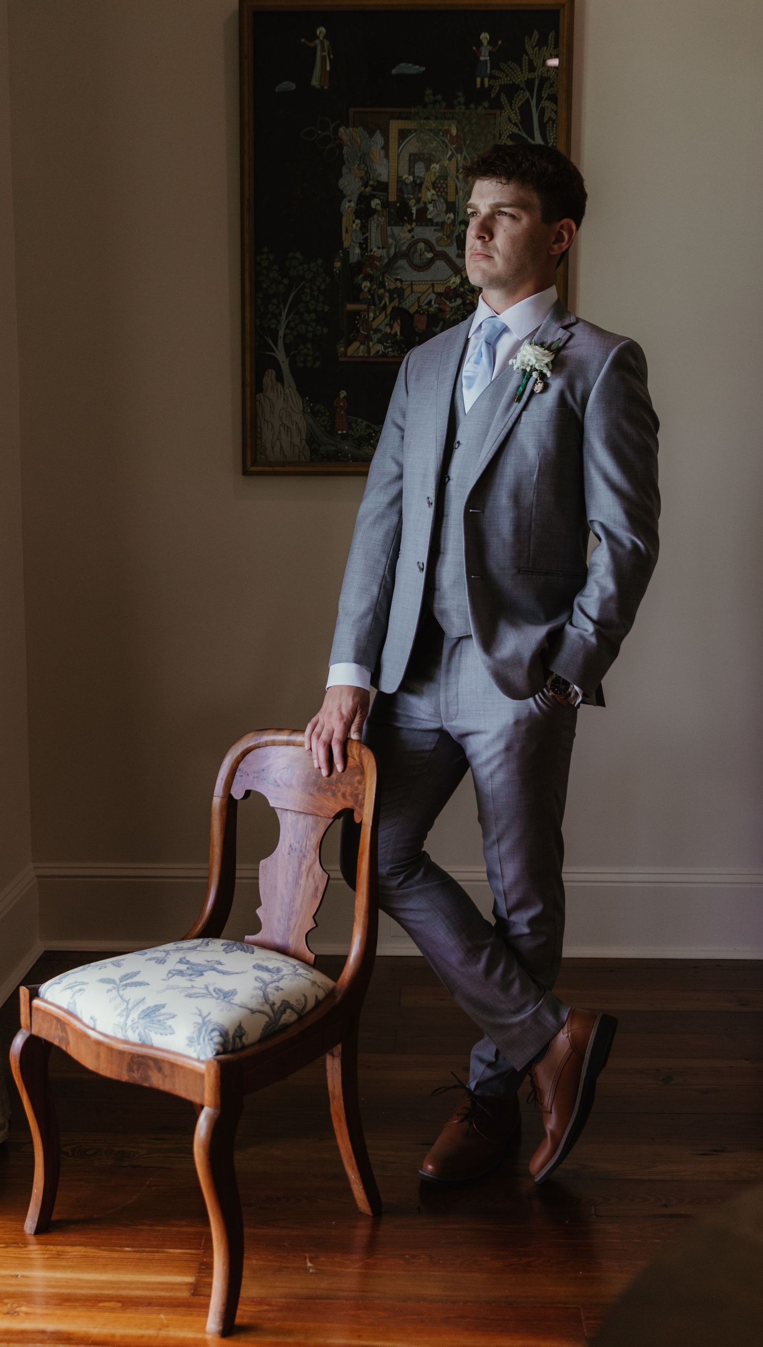 Formal portrait of someone in a gray suit and boutonniere leaning against an antique chair in moody lighting.