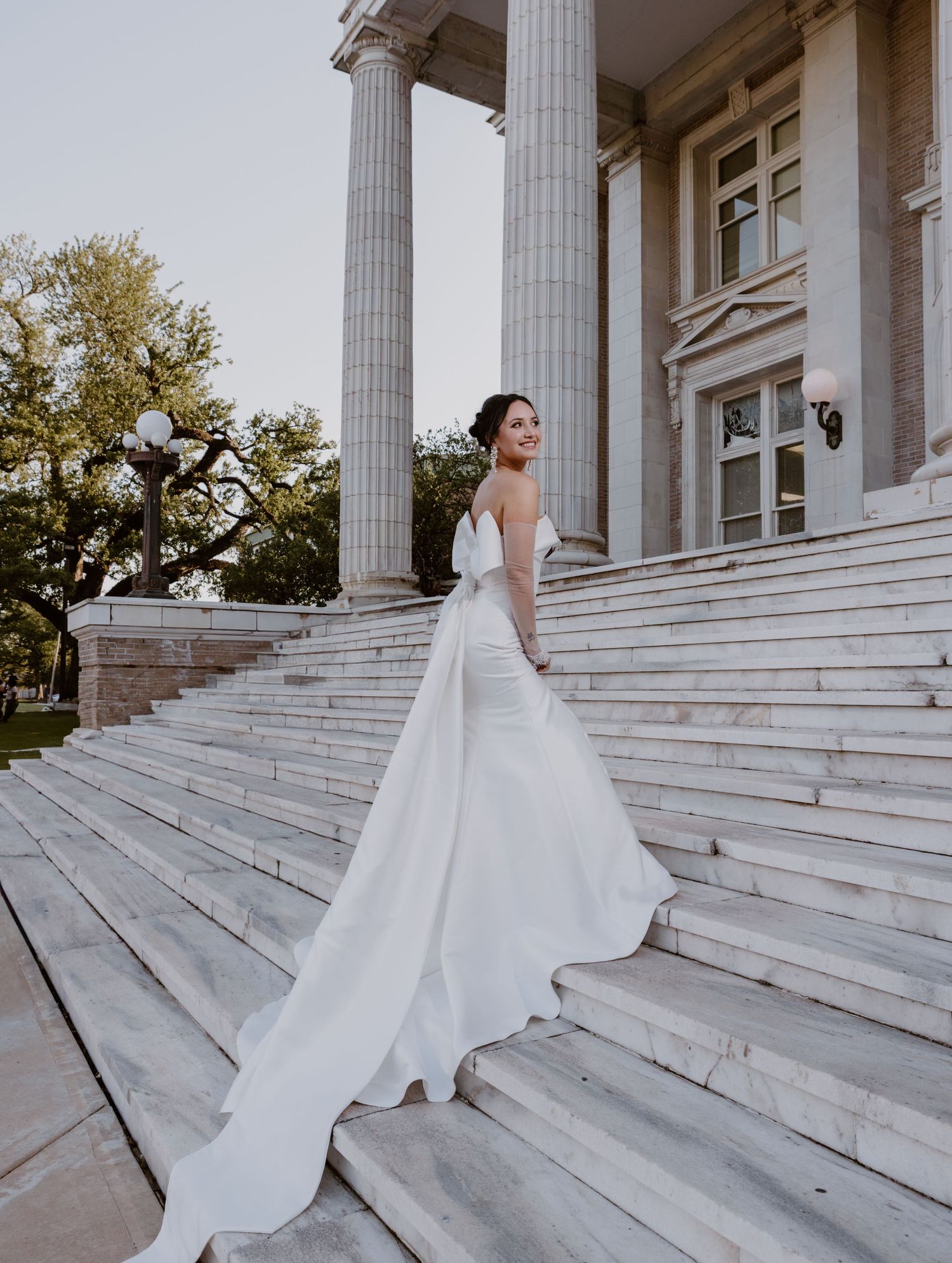 A bride in a flowing white wedding dress stands gracefully on grand marble steps of a classical building with pillars.