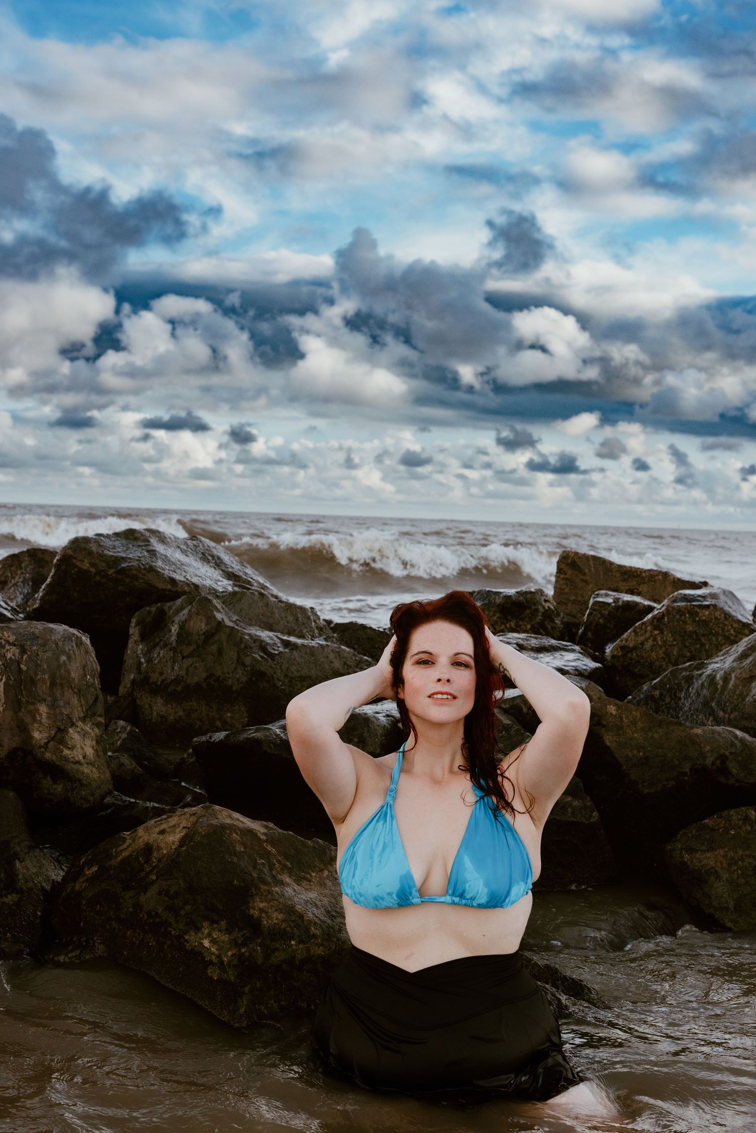 A person in a turquoise bikini top poses among rocky coastal formations under dramatic cloudy skies at the beach.