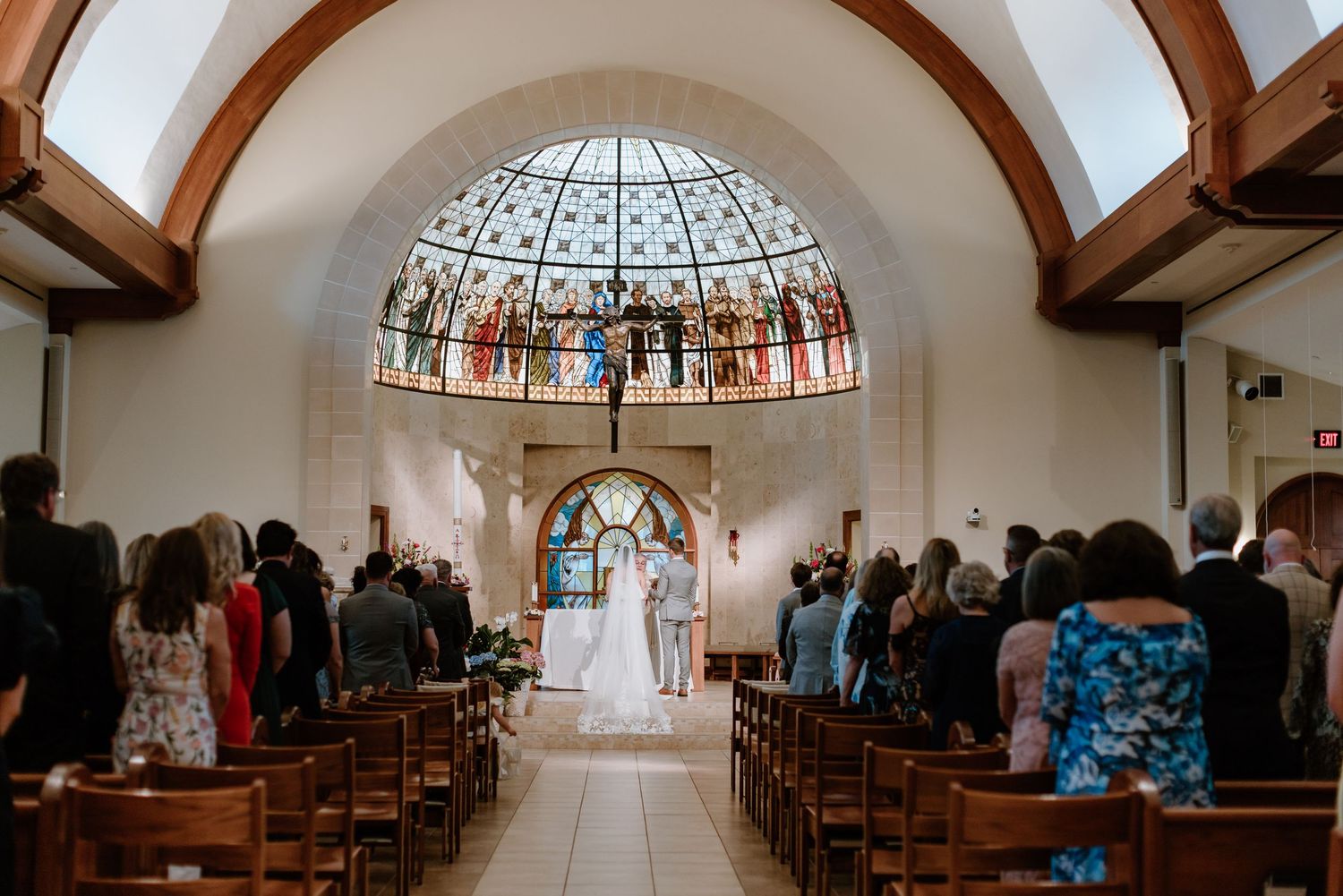 A wedding ceremony in a beautiful chapel with a stained glass dome ceiling and wooden pews filled with guests.