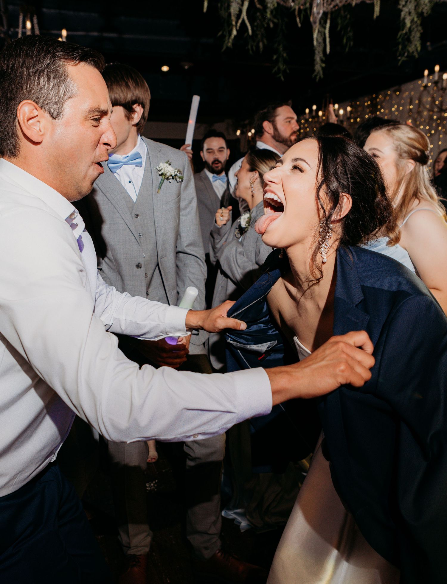 Wedding guests sing and dance energetically during a lively reception party under twinkle lights.