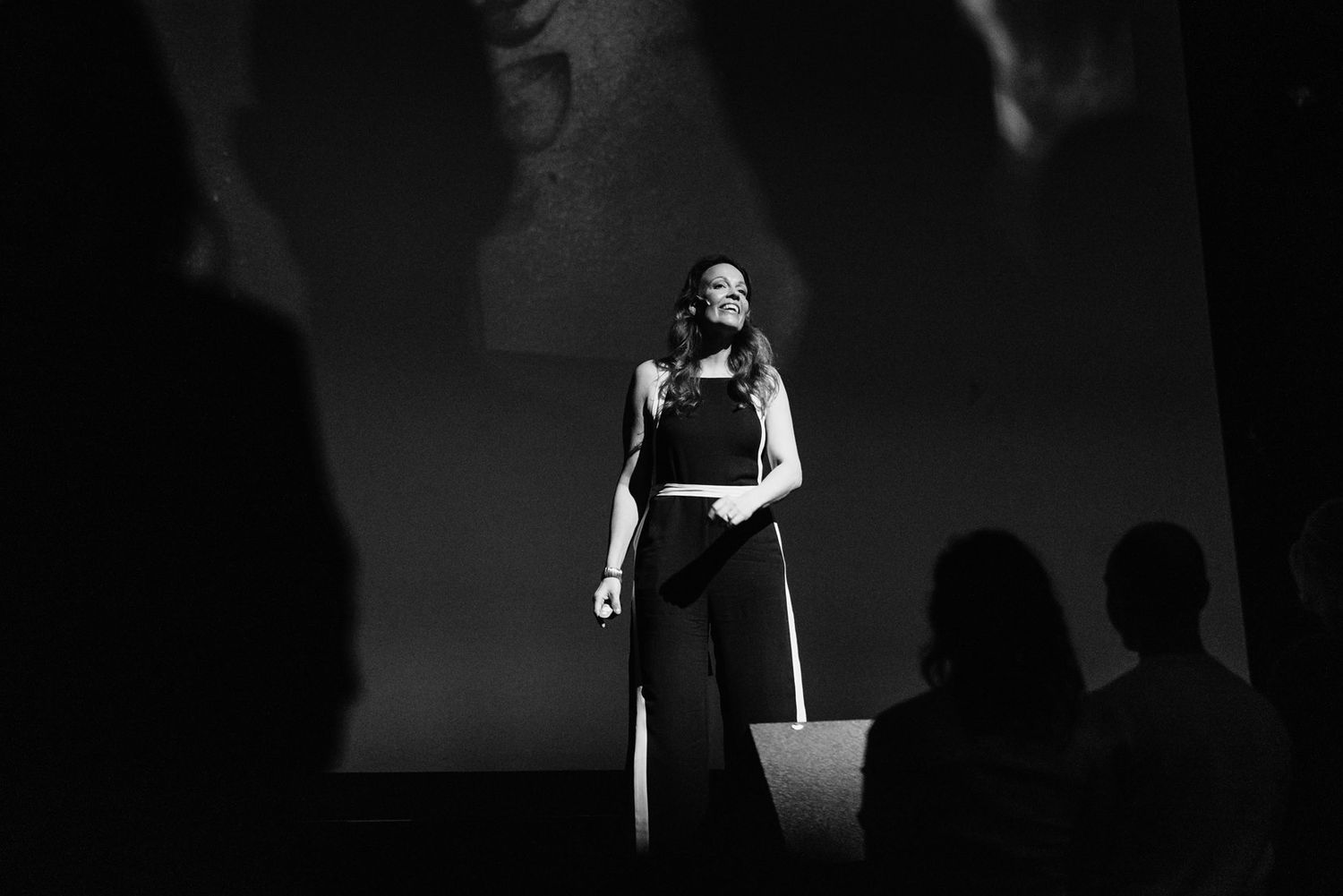 A performer stands on stage in a black and white photograph with silhouetted audience members visible in the foreground.