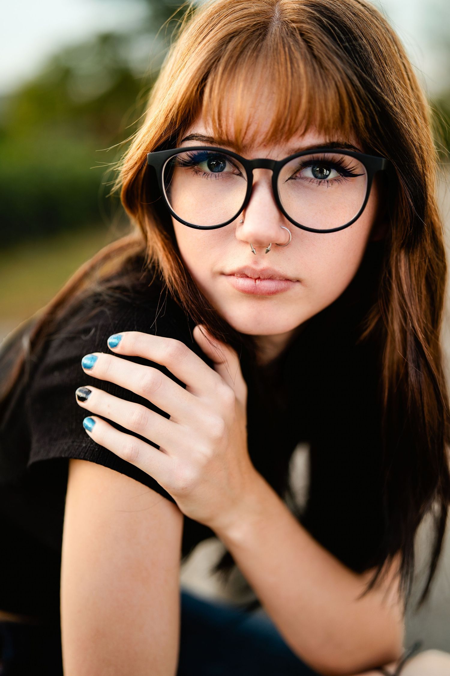 Portrait of someone wearing black-framed glasses and dark clothing with blue nail polish visible on their hand.