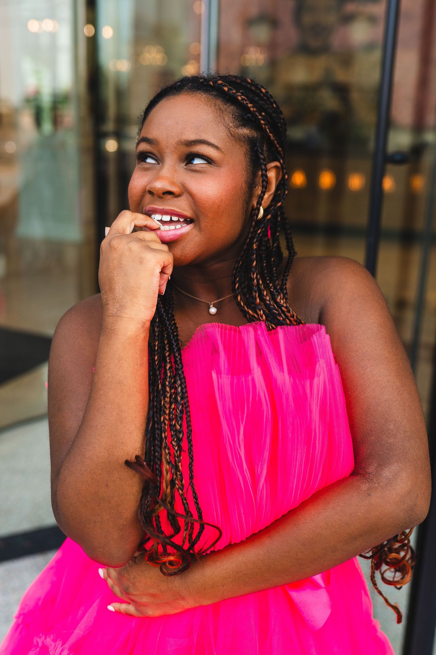 Smiling woman in vibrant pink dress with braided hair looks off to the side while resting chin on hand.