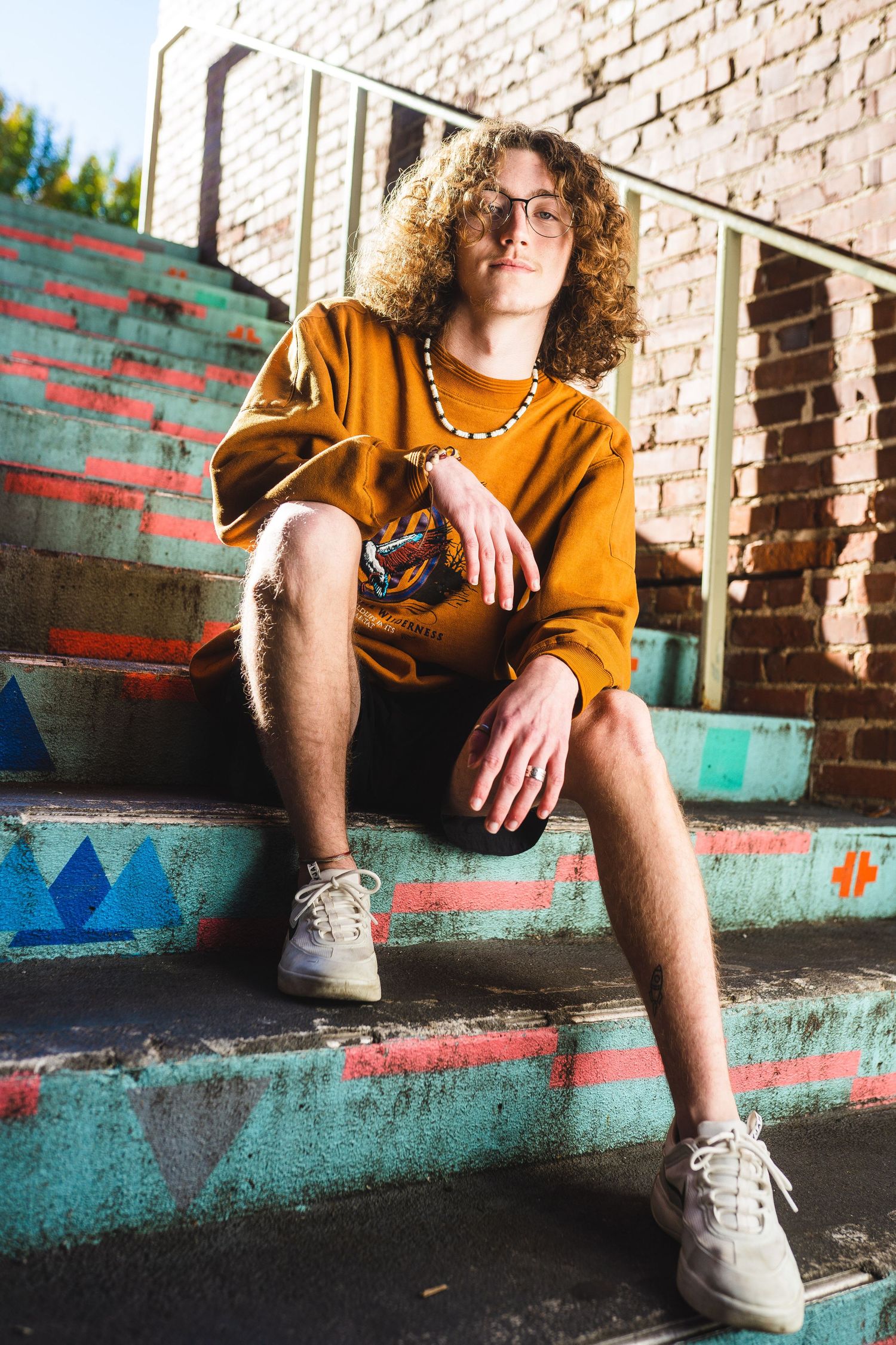 Person in orange sweatshirt and shorts sits on colorful painted concrete steps against brick wall wearing white sneakers.