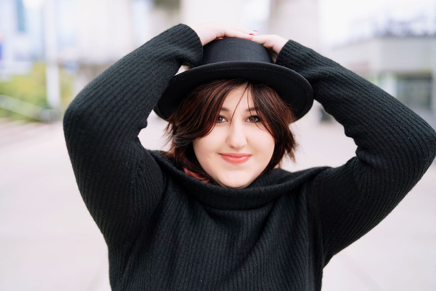 A person in a black turtleneck sweater and dark hat smiles while holding their hands up to their head outdoors.