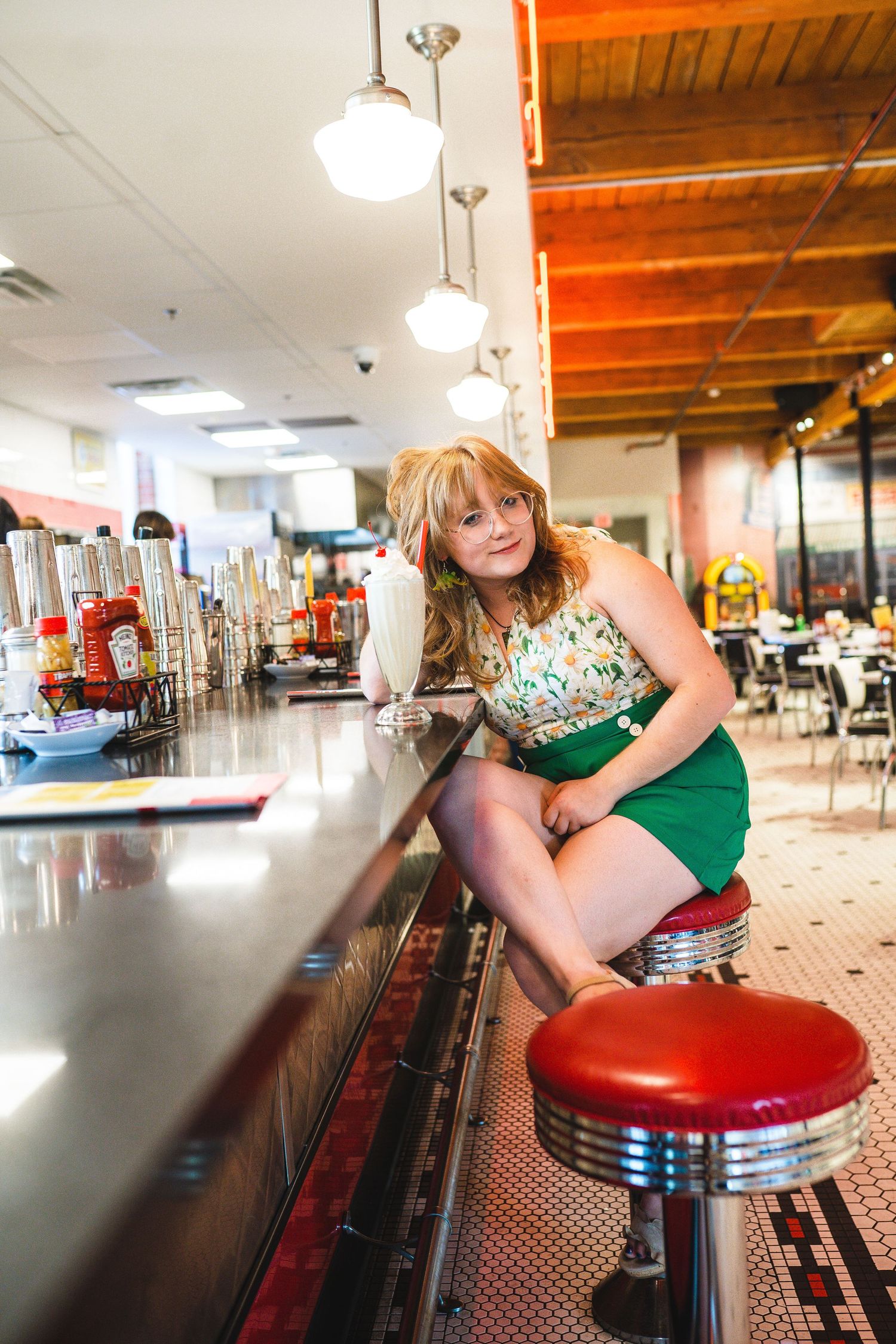 Person in vintage-inspired outfit sitting on red diner stool at retro counter under globe lights in classic diner setting.