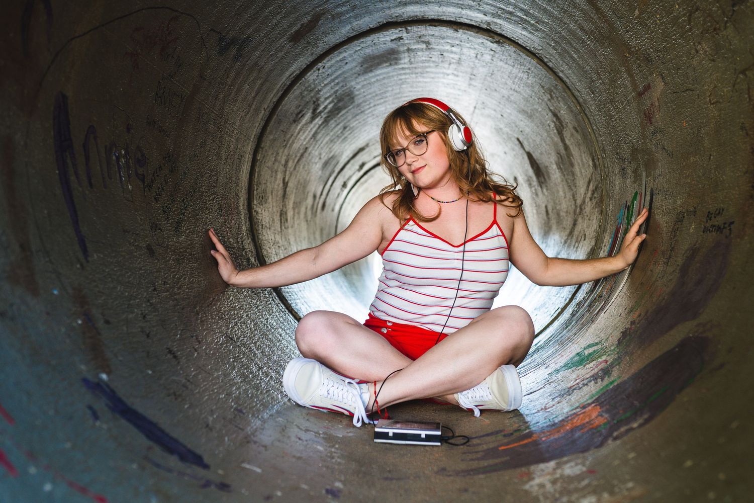 Person in striped top and shorts sits cross-legged inside large concrete drainage tunnel while touching the walls.