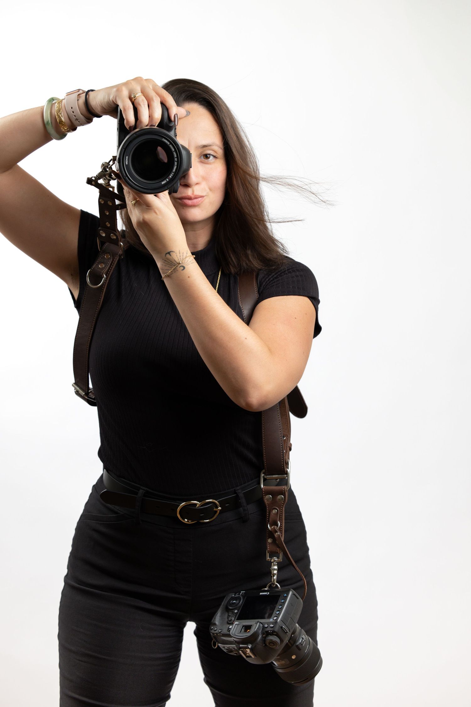 A photographer in all black clothing holds up a professional camera against a white background.