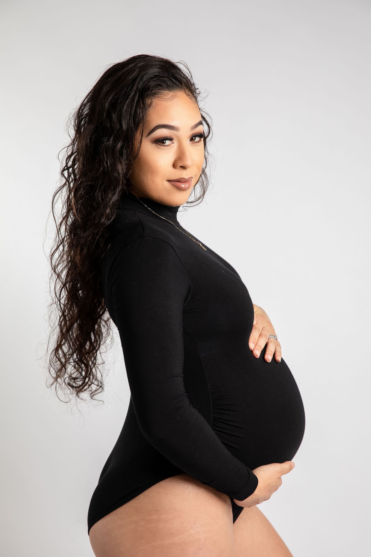 Side view maternity photo of person in black bodysuit showing pregnant belly against white backdrop.
