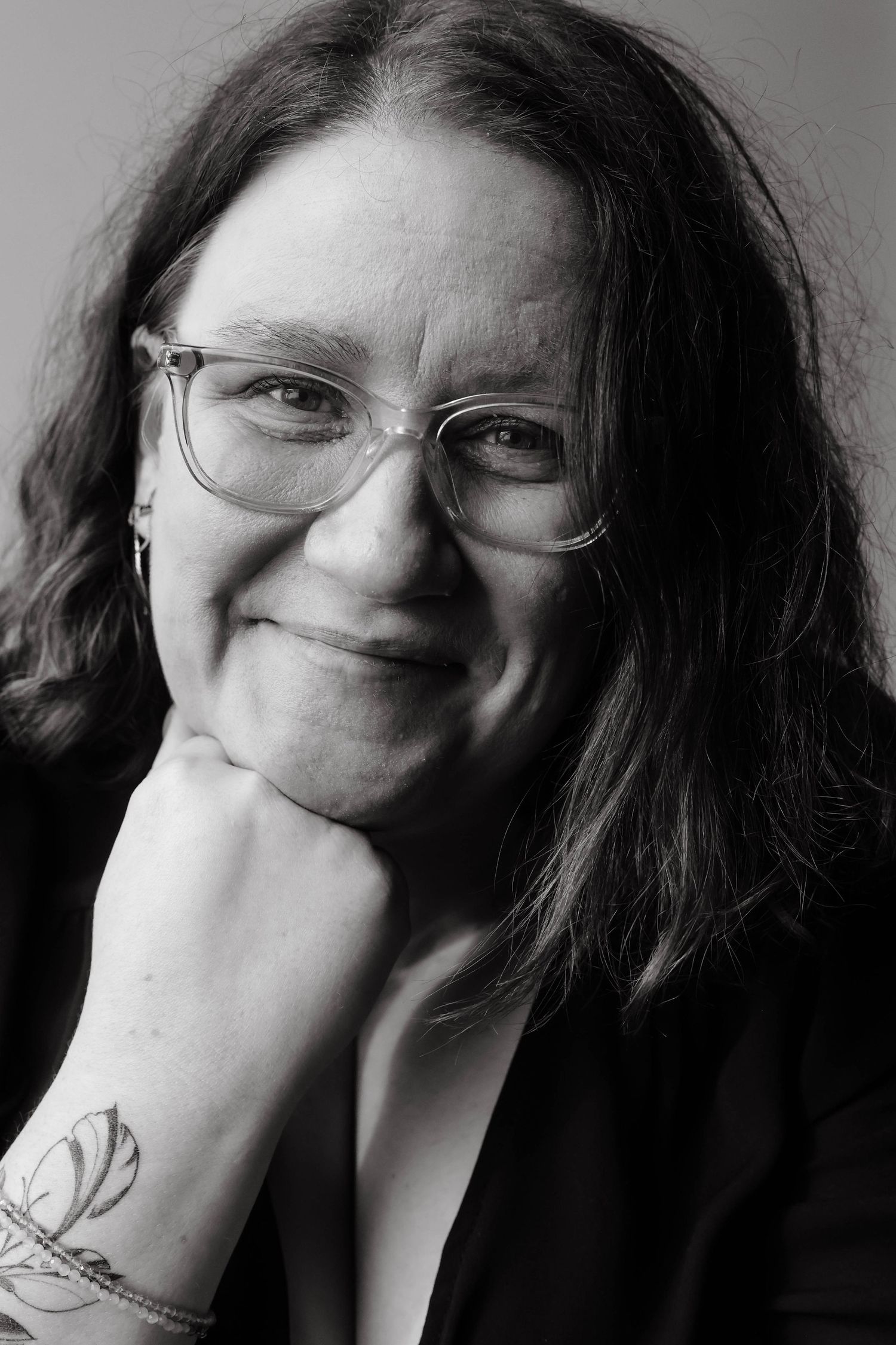 Black and white portrait showing a person with glasses and long dark hair smiling warmly while resting their tattooed hand on their chin.