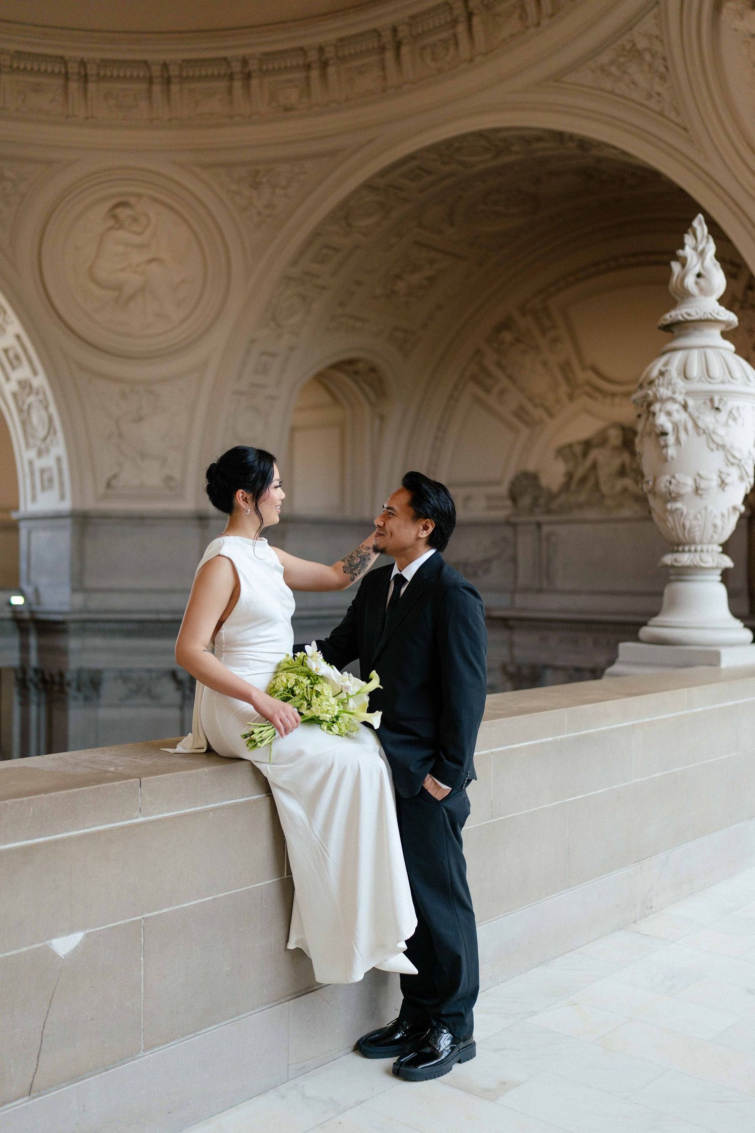 Newlywed couple shares intimate moment in elegant hallway with ornate architectural details and classical decorative urn.