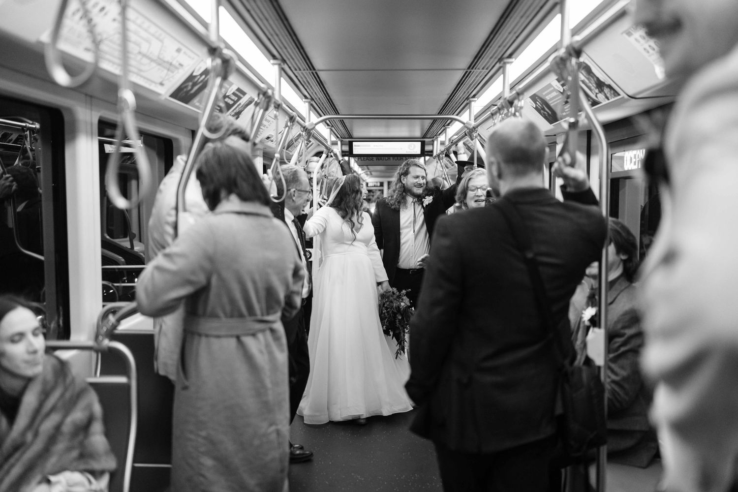 A bride in a white wedding dress stands in a crowded subway car among commuters in black and white photograph.