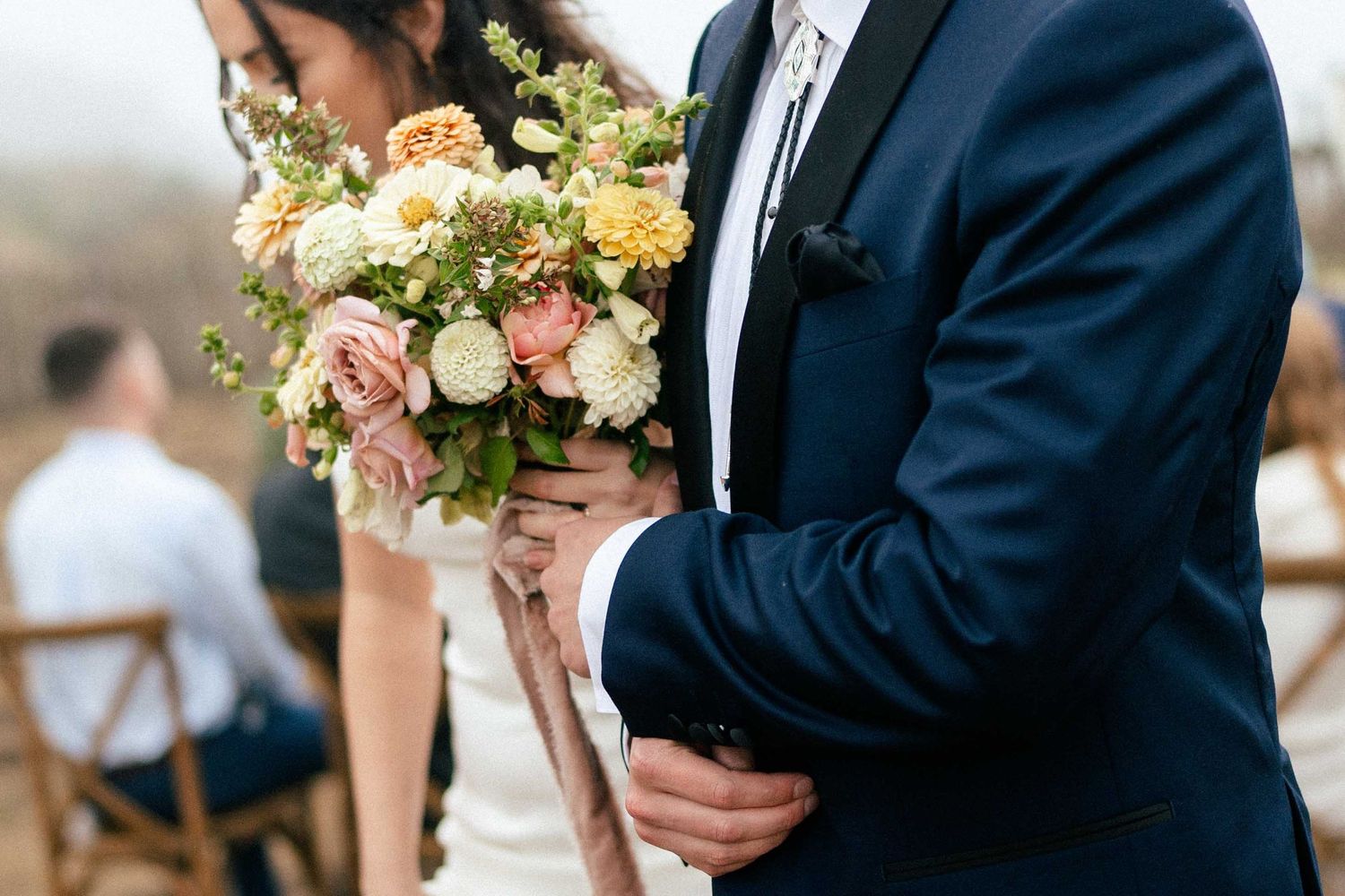 A wedding couple holds a beautiful bouquet of pink and cream flowers with their arms linked, wearing a navy blue suit.