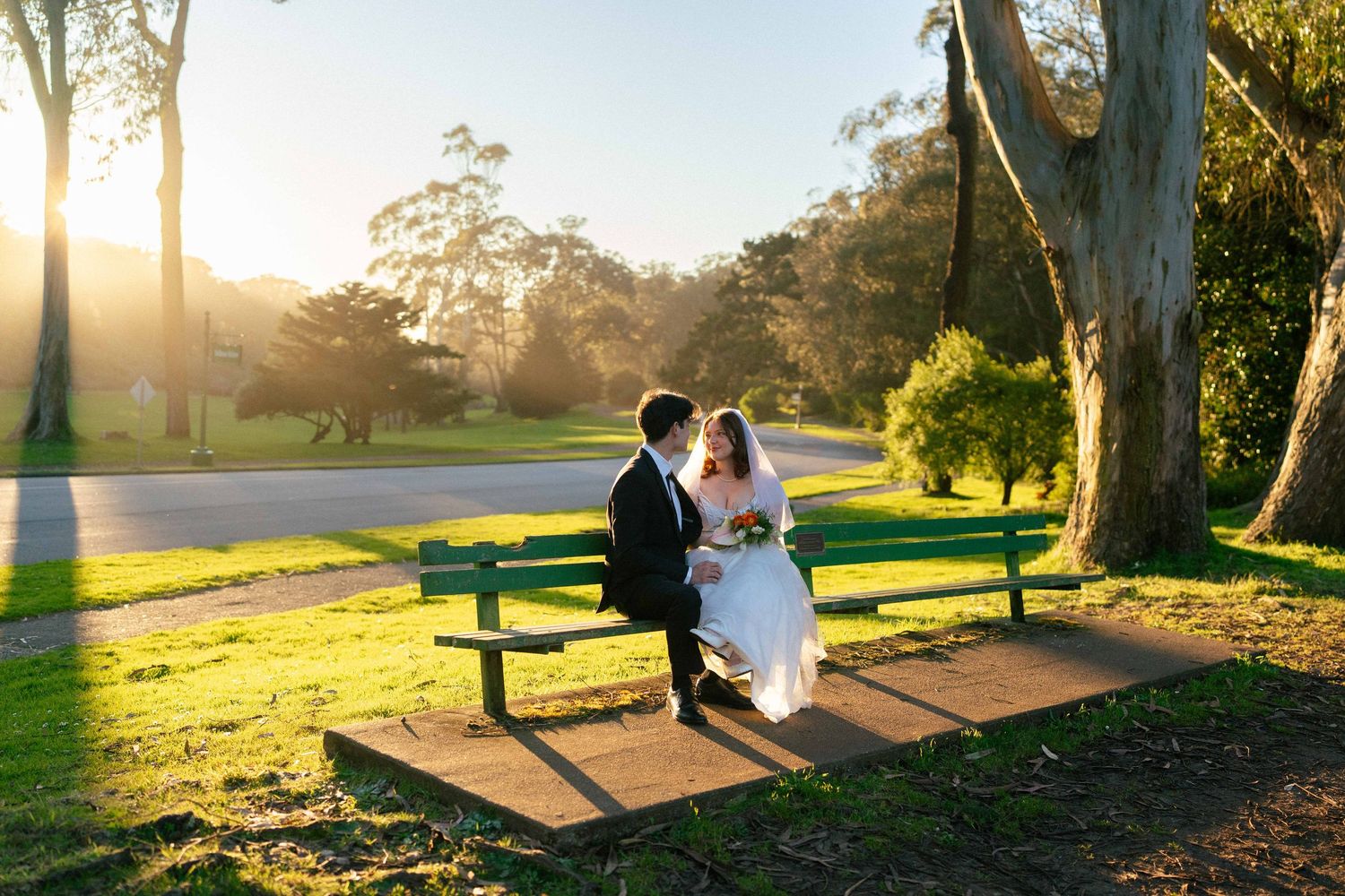 Couple in wedding attire sits together on park bench during golden sunset in lush outdoor setting.