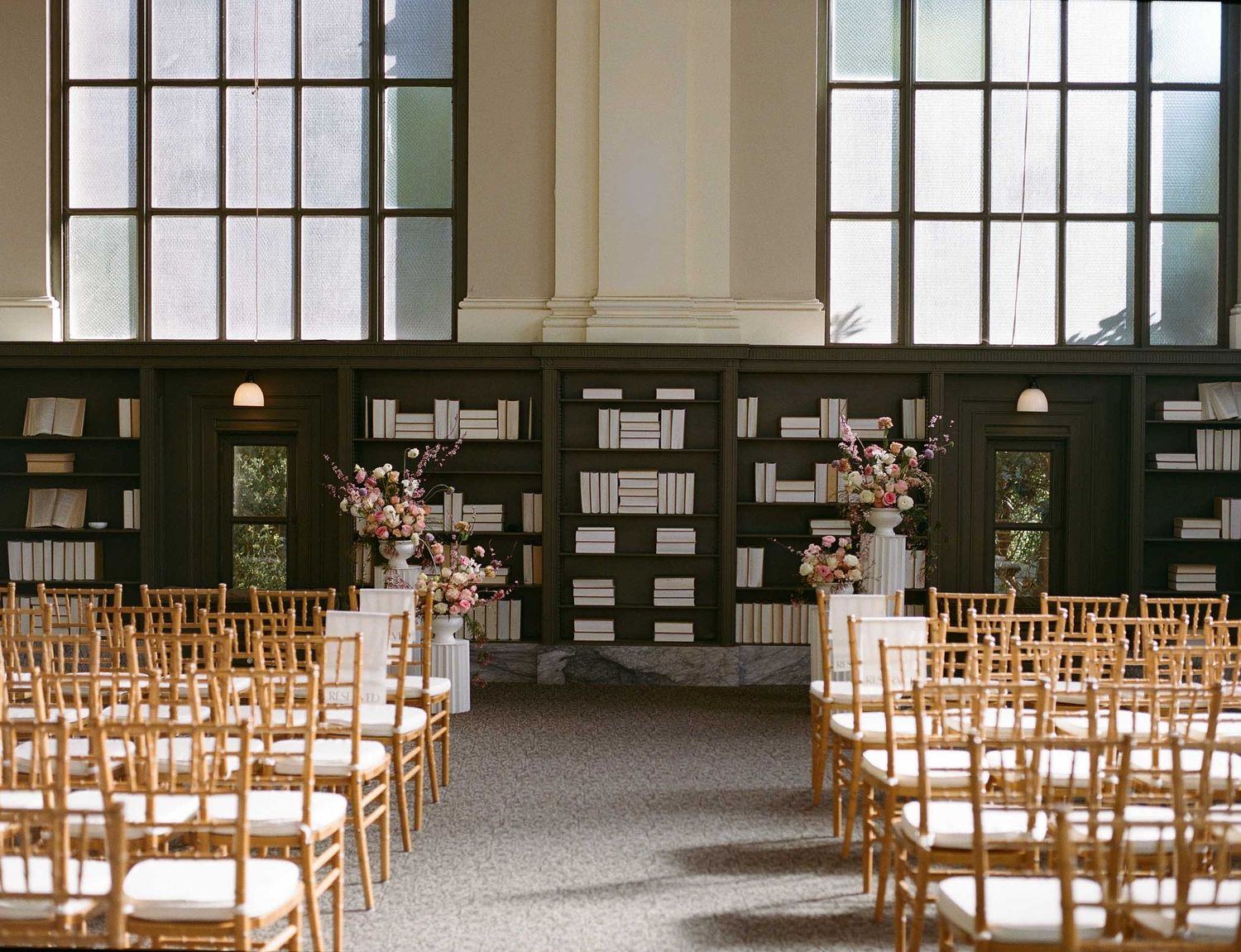 Elegant wedding ceremony setup with wooden chiavari chairs facing wall-mounted bookshelves decorated with floral arrangements.