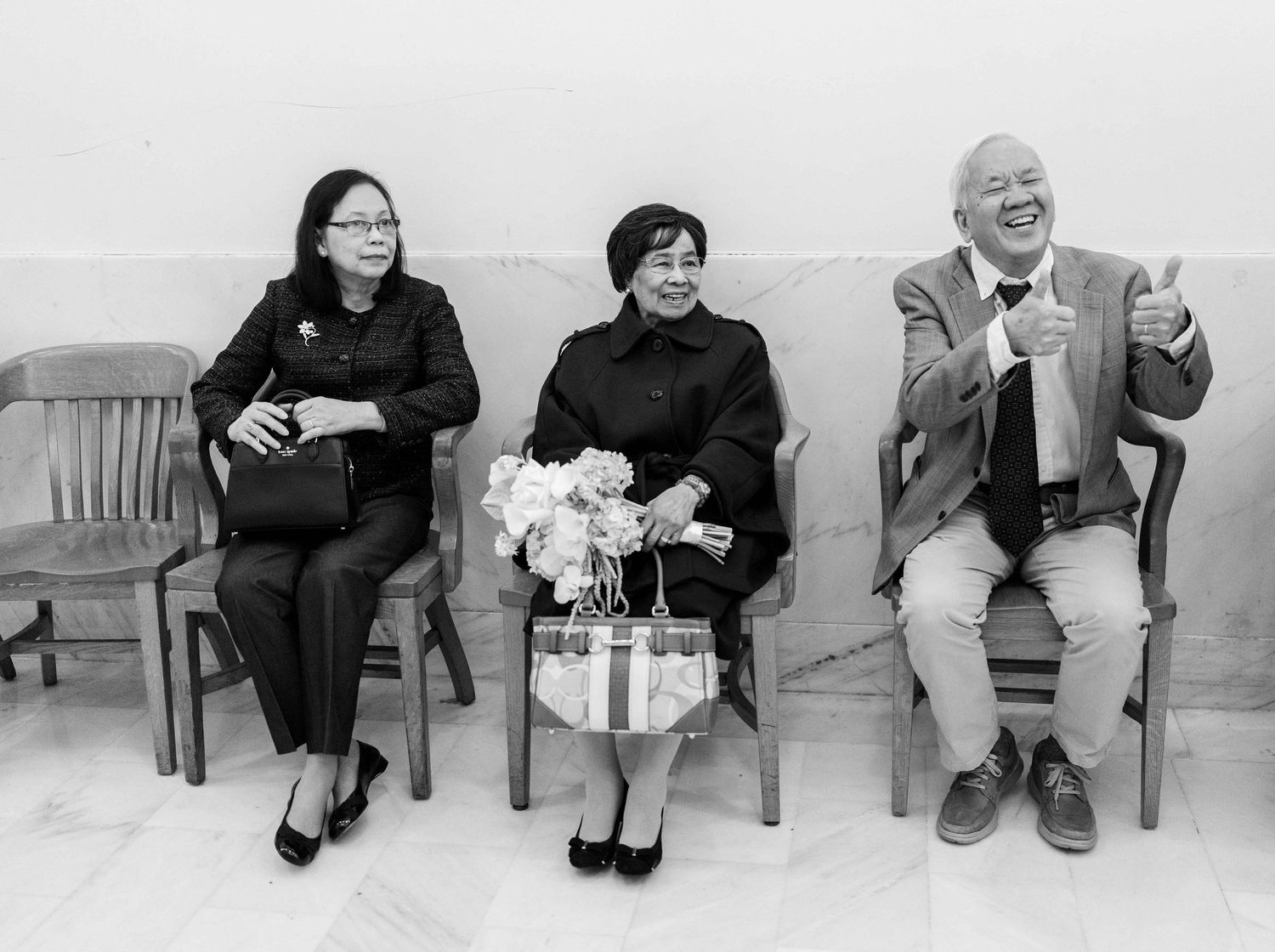 Three people sit on wooden chairs against a white wall in a black and white photograph.