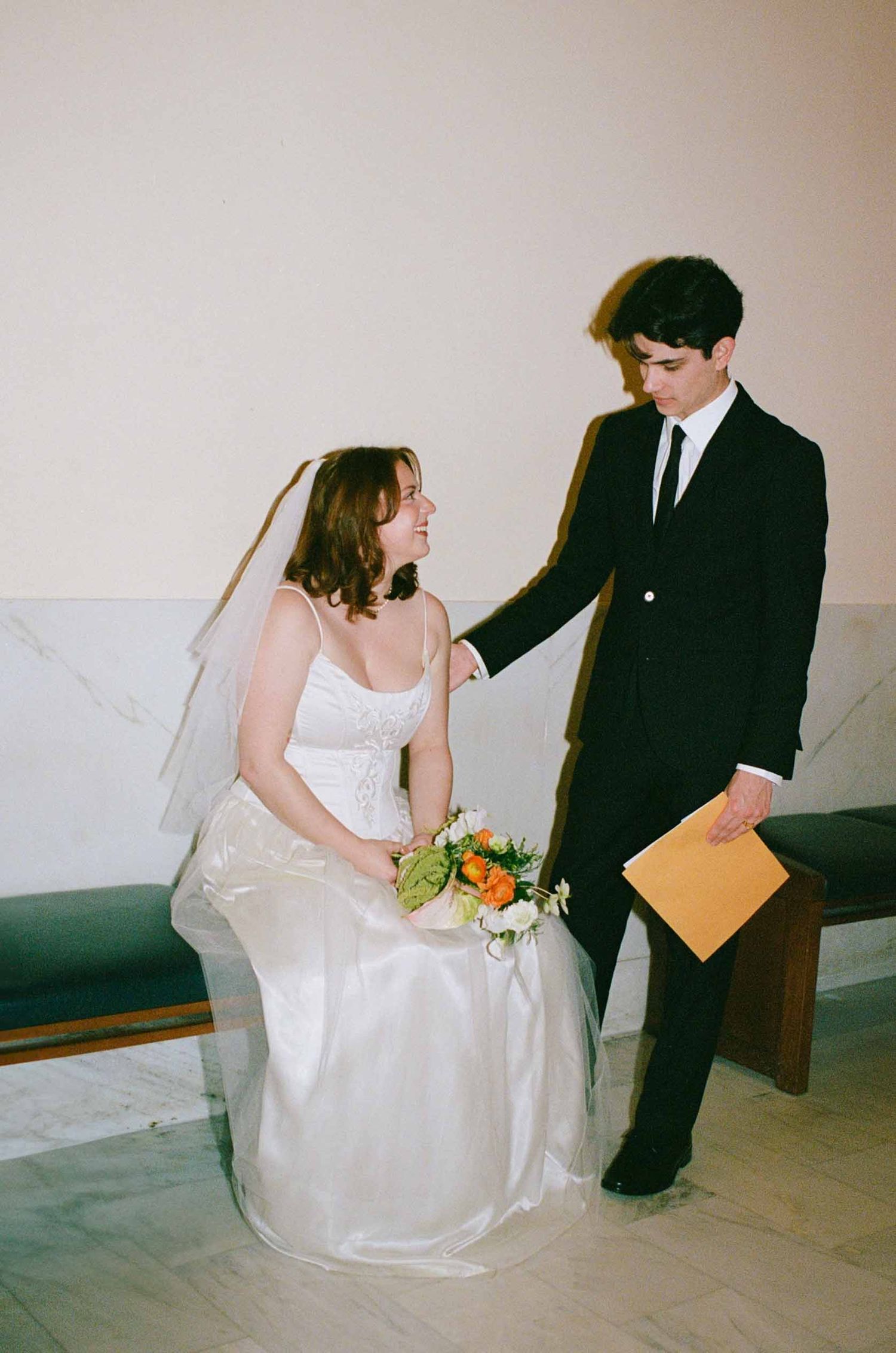 A tender moment captured at a wedding ceremony showing two figures in a formal setting with one seated in white.