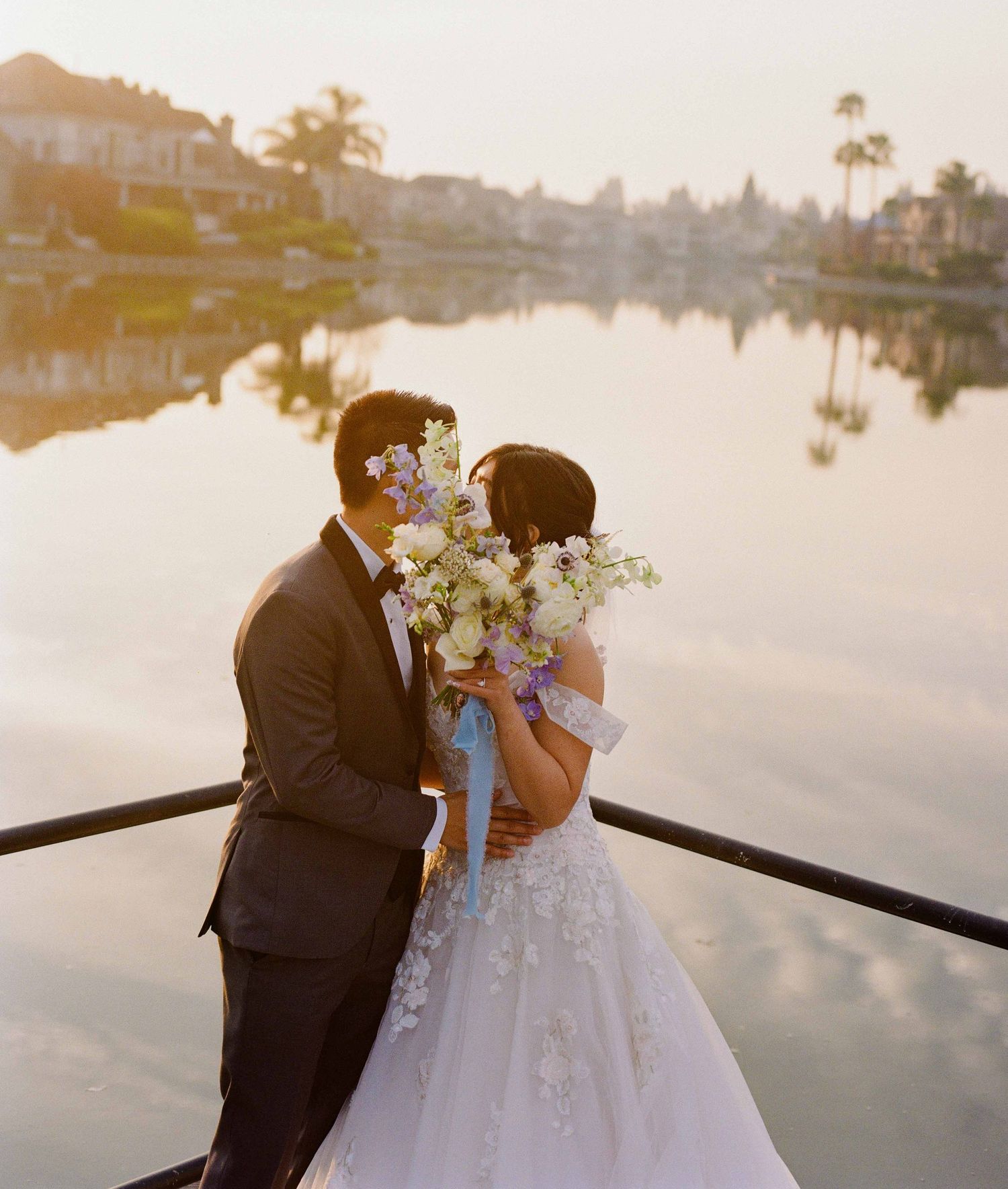 A couple shares a romantic moment on a boat at sunset beside a tranquil lake with white floral bouquet and wedding attire.