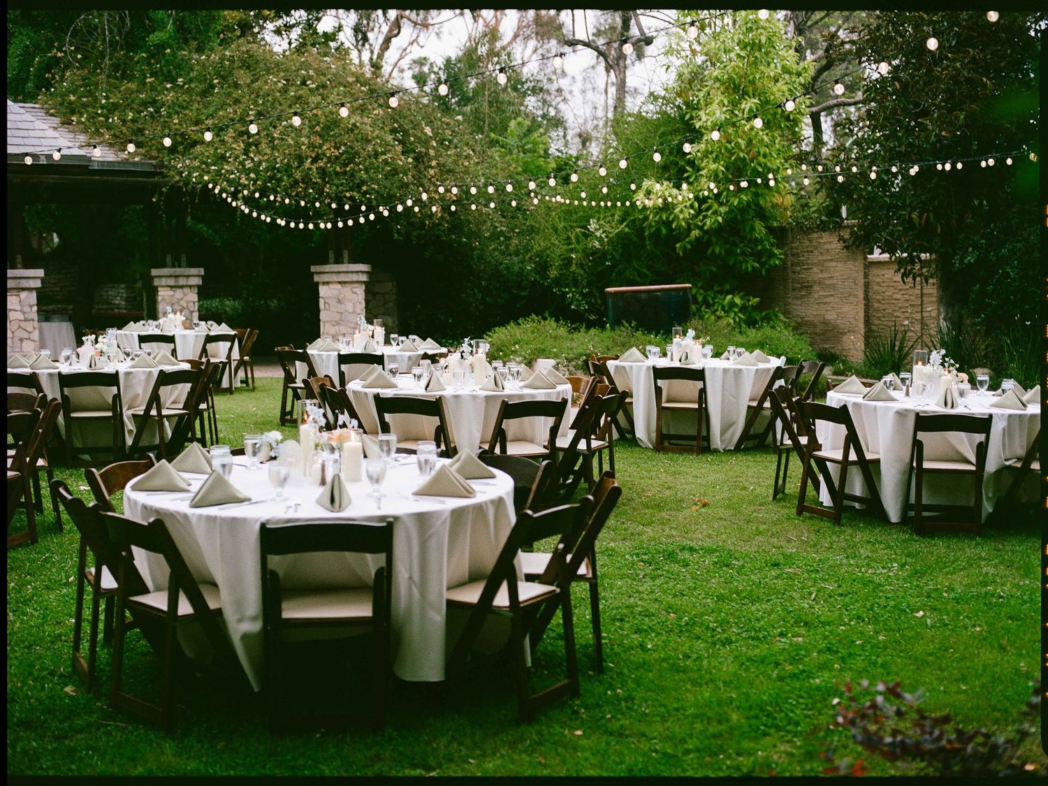 An outdoor garden reception setup with round tables, white linens, and string lights creating a romantic atmosphere.