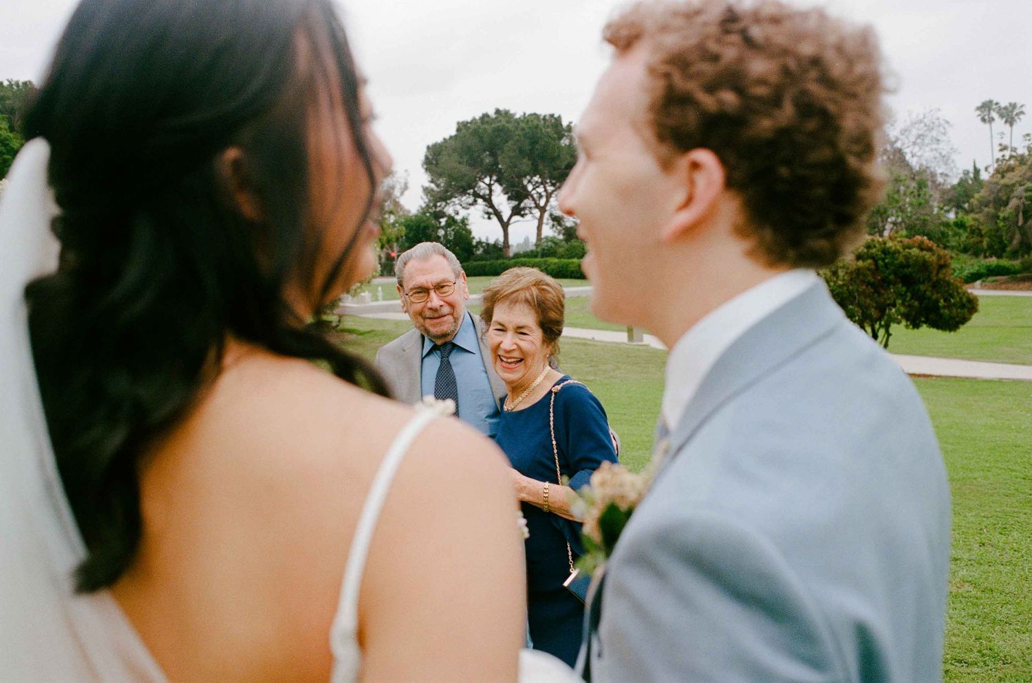 A candid moment at an outdoor wedding ceremony with people interacting on a grassy lawn under trees.