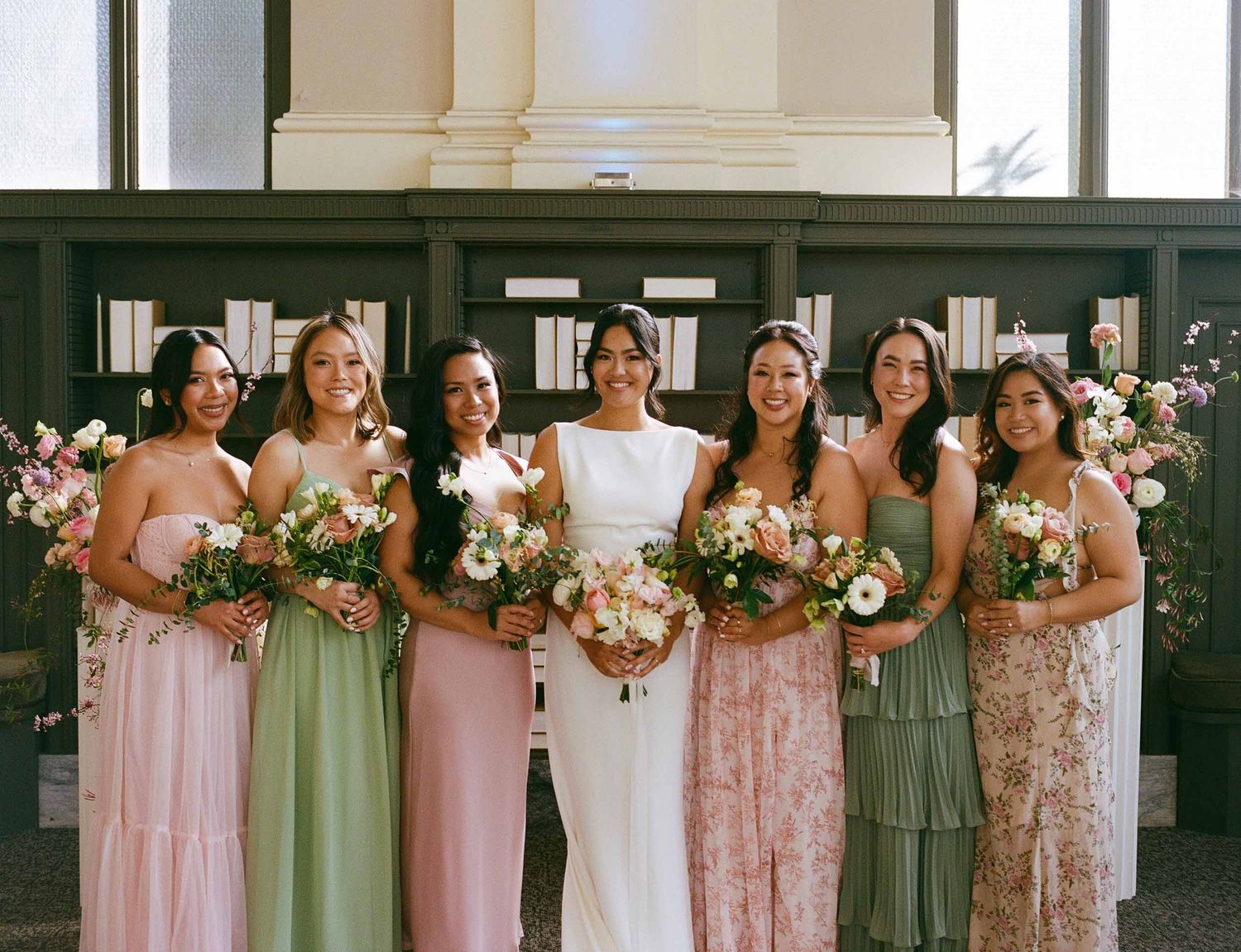 Bridesmaids in pastel pink and sage green dresses stand together holding floral bouquets at a wedding ceremony.