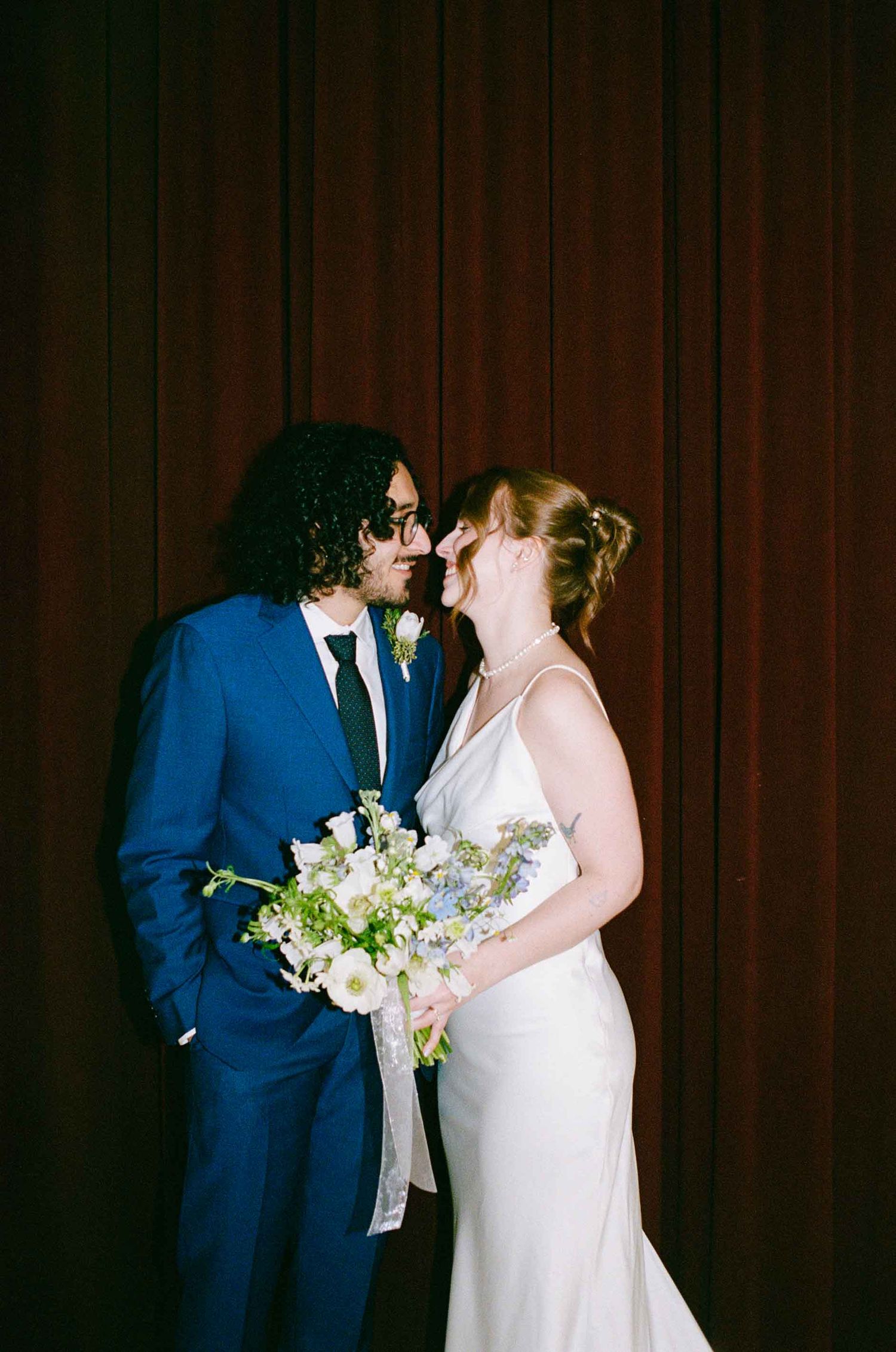 A couple shares a romantic moment on their wedding day, with the bride holding a delicate white and blue floral bouquet.