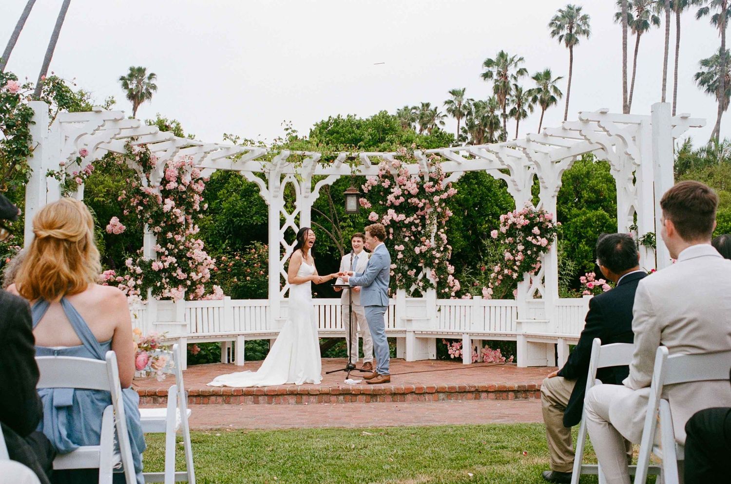 An intimate outdoor wedding ceremony takes place under a white lattice arbor adorned with flowers and palm trees.