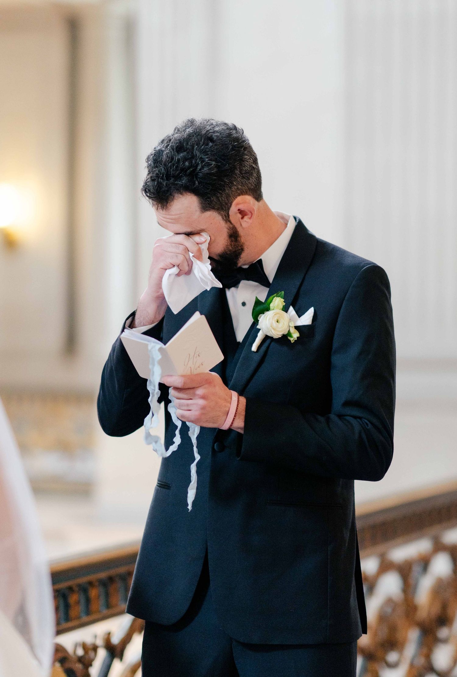 An emotional moment captured during a wedding ceremony as a groom in a black tuxedo reads a note while wiping tears.