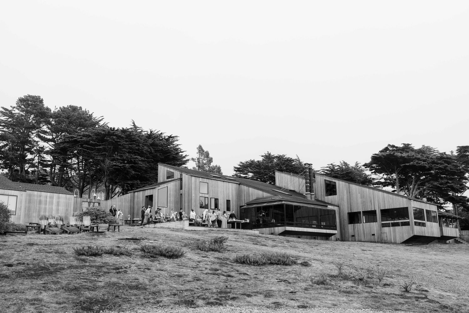 Black and white photograph of simple military-style housing units with people gathered outside near pine trees.