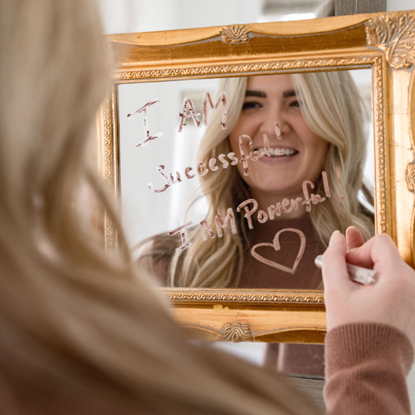 Person writing 'Success is Possible' with a heart symbol on a mirror in an ornate gold frame.