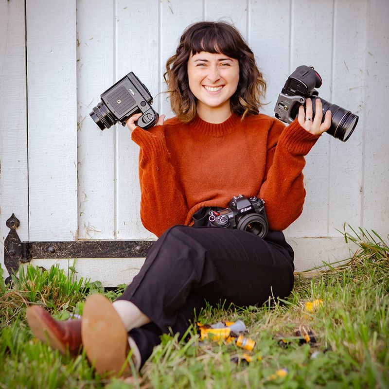 A photographer in an orange sweater displays camera equipment while sitting on grass.