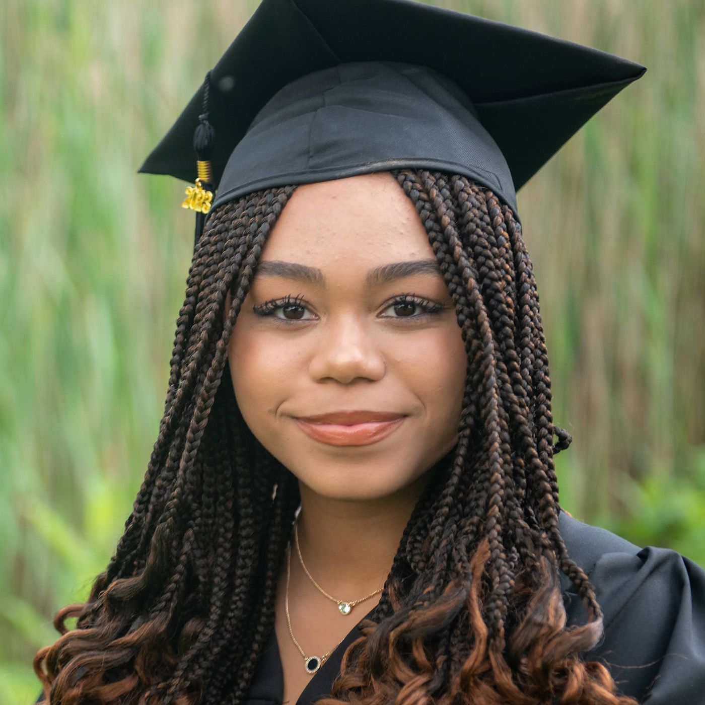 Graduate in traditional black cap and gown with long braided hair poses outdoors.