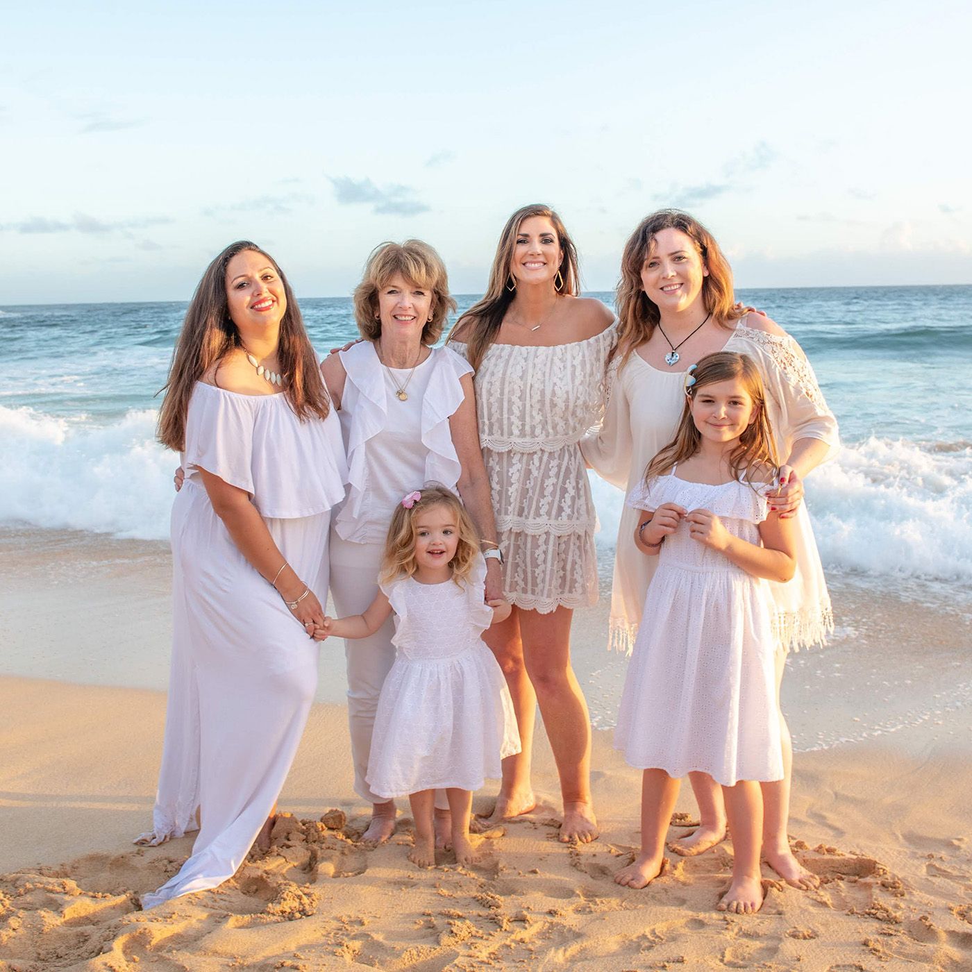 Group portrait on beach at sunset wearing white dresses with ocean waves in background.