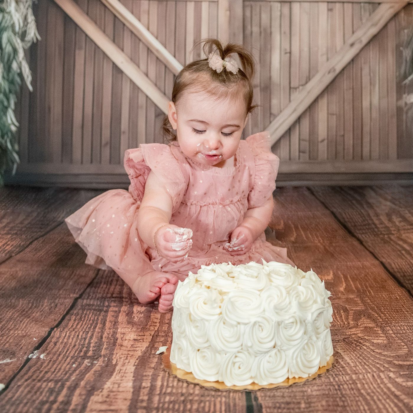 Baby in pink dress reaches for white frosted cake on wooden floor.